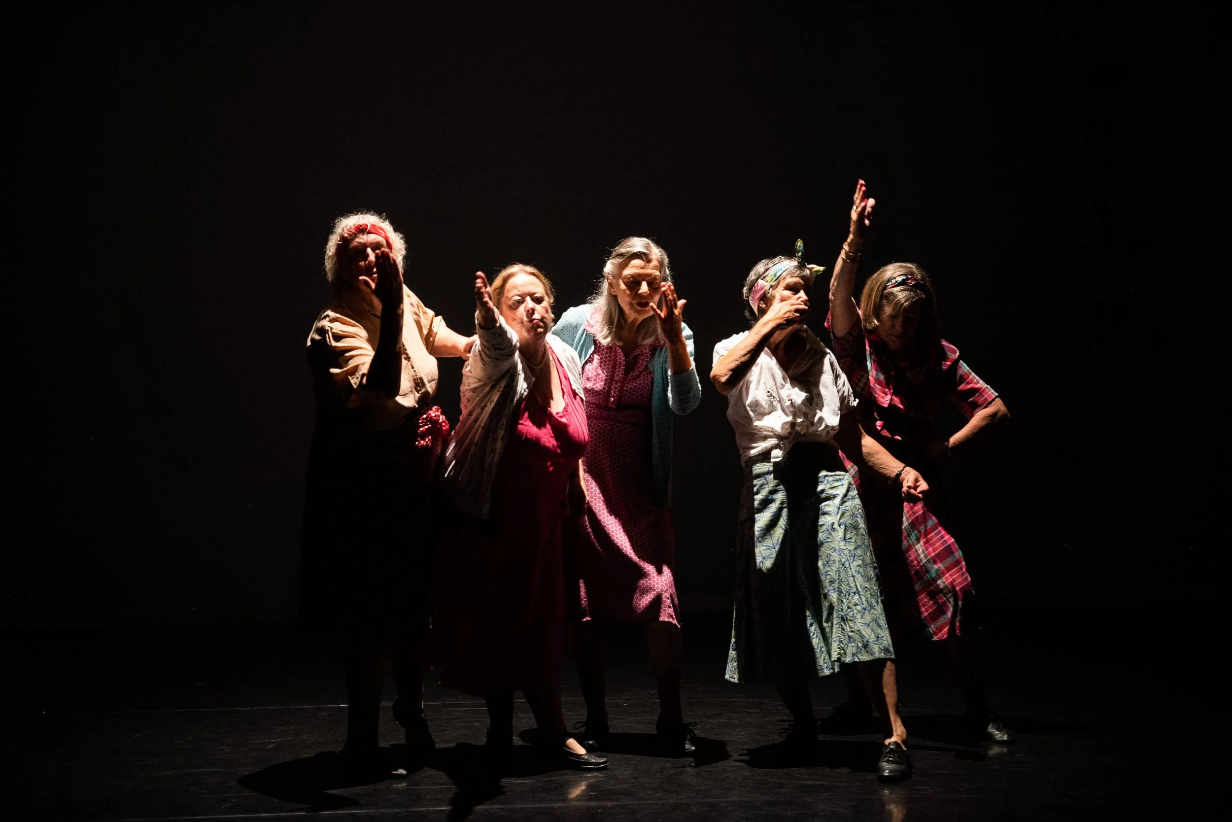 Five older women performers in colourful everyday clothes gesture with raised hands and arms on a darkly lit performance stage.