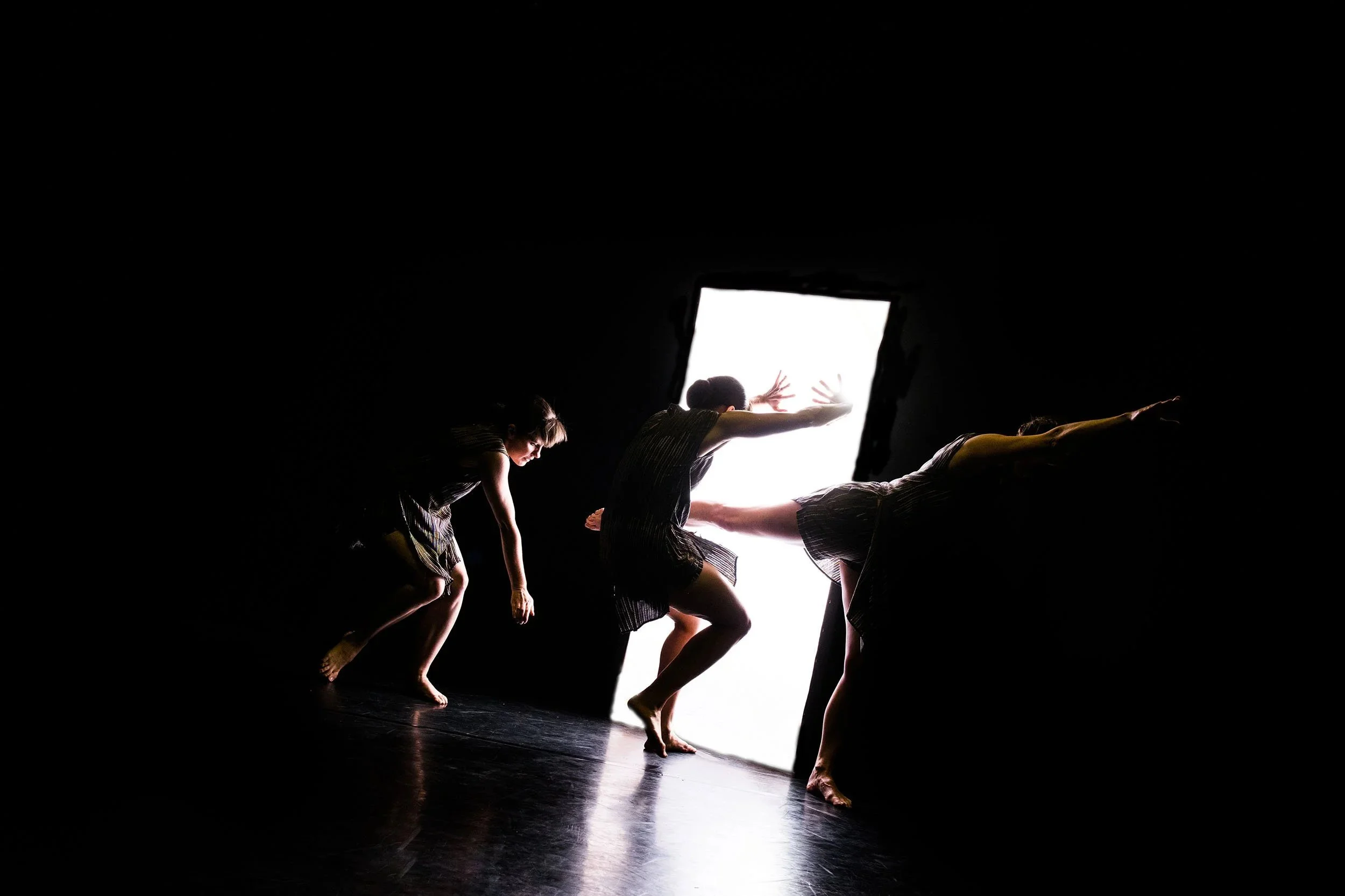 Contemporary dancers moving toward a bright light portal in a dark stage performance by NOCTURN, directed by John Darvell.