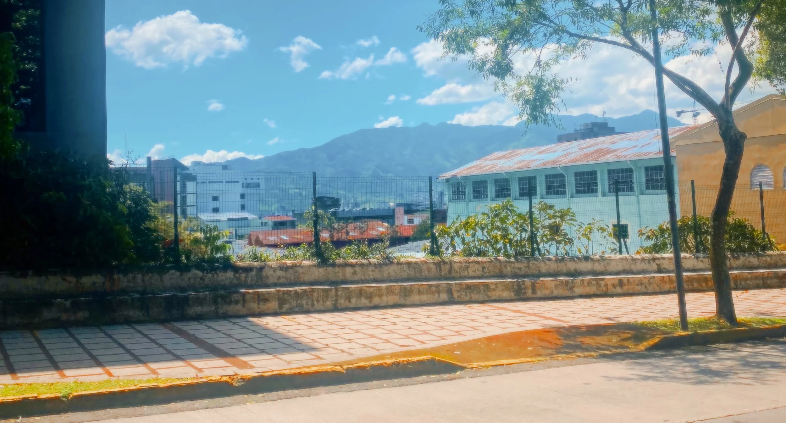 View of a cityscape with buildings, trees, a fence, and mountains in the background under a partly cloudy blue sky.
