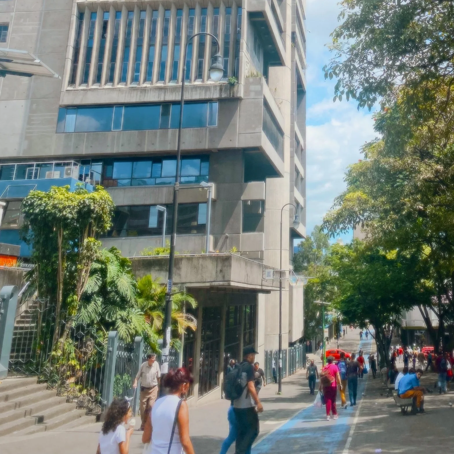A busy city street with pedestrians walking, tall modern buildings, and large trees providing shade, under a partly cloudy sky.