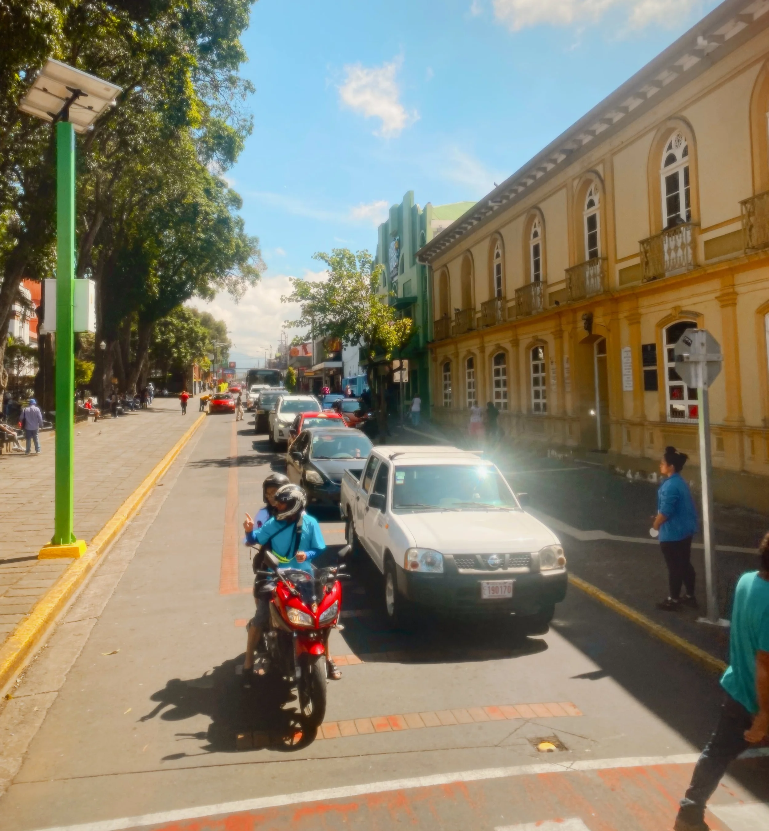 Street scene with yellow historic building, green and white buildings, trees, cars parked along the street, pedestrians walking, a motorcycle with a rider wearing a helmet and blue jacket, and a person standing beside the motorcycle.