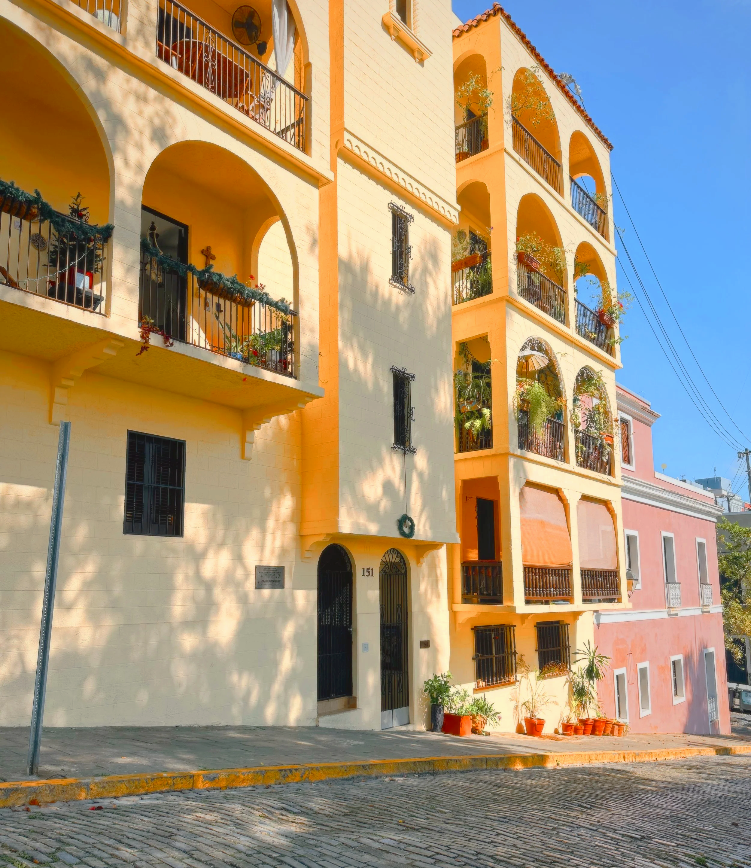 A multi-story building with arched balconies, decorated with potted plants and a wreath, painted in shades of yellow and pink, under a clear blue sky, with a cobblestone street in front.