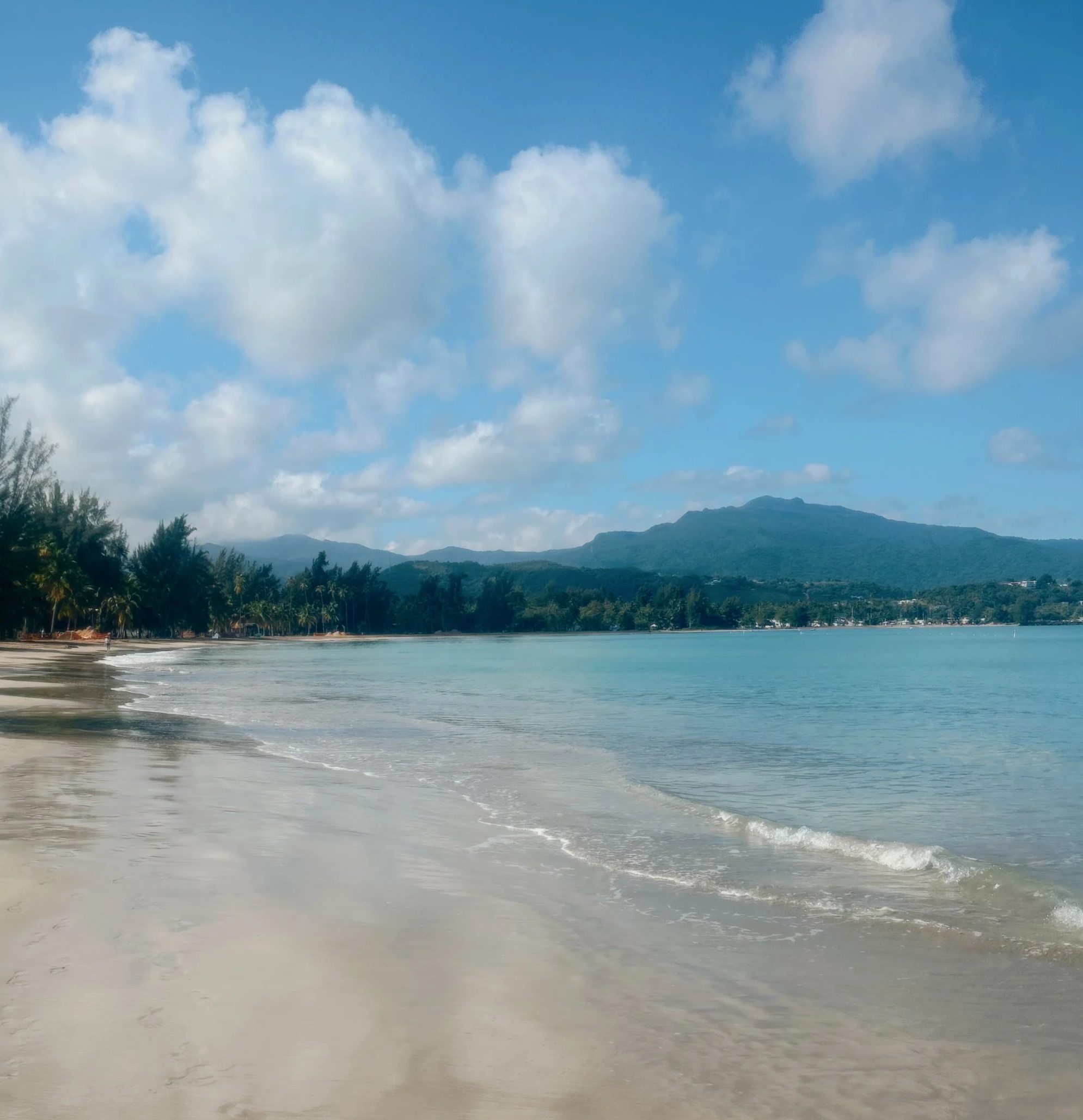 A tropical beach with white sand, palm trees, blue sky with clouds, and a mountain in the background.