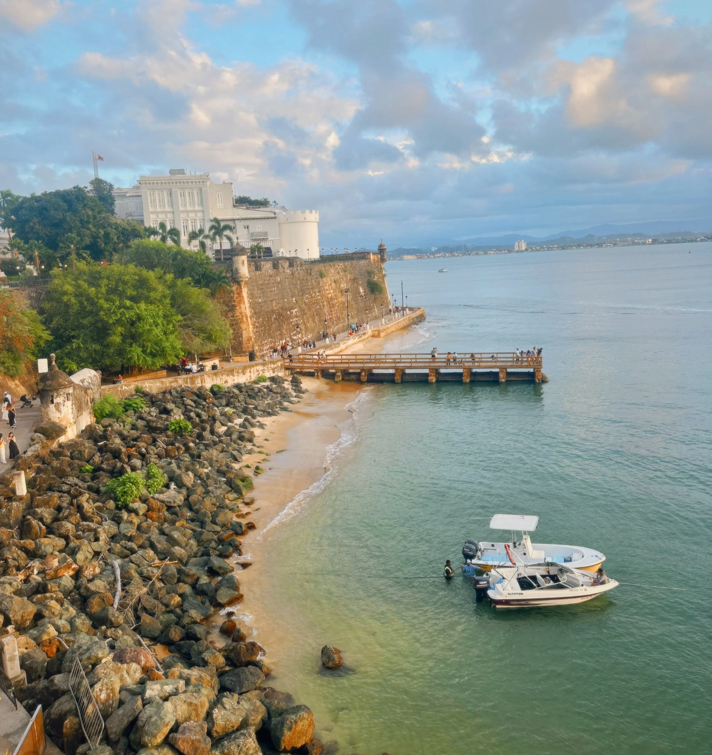View of a historic coastal fortress with white buildings on a cliff, a promenade with people walking, and boats docked in the water on a cloudy day with blue sky.
