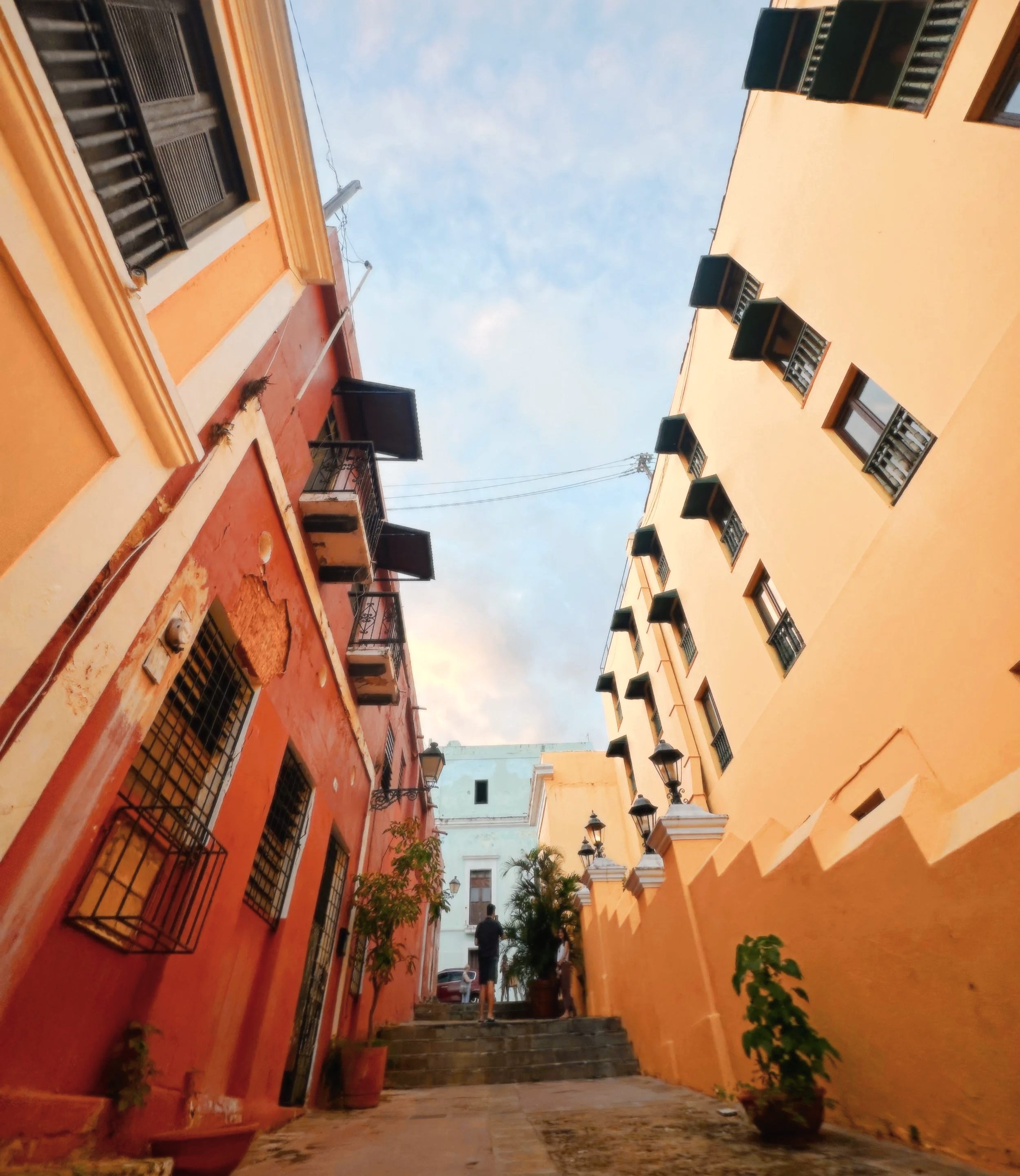 Narrow courtyard with colorful walls, potted plants, lanterns, and a person walking up steps, under a partly cloudy sky.