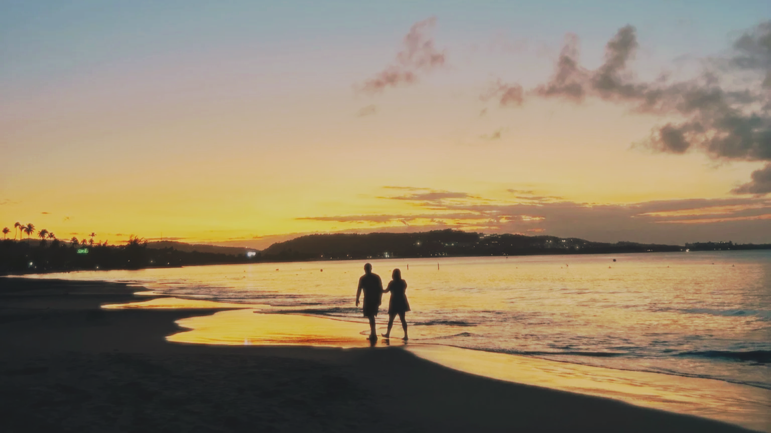 A couple holding hands walking along the shoreline at sunset, with a colorful sky and calm water.