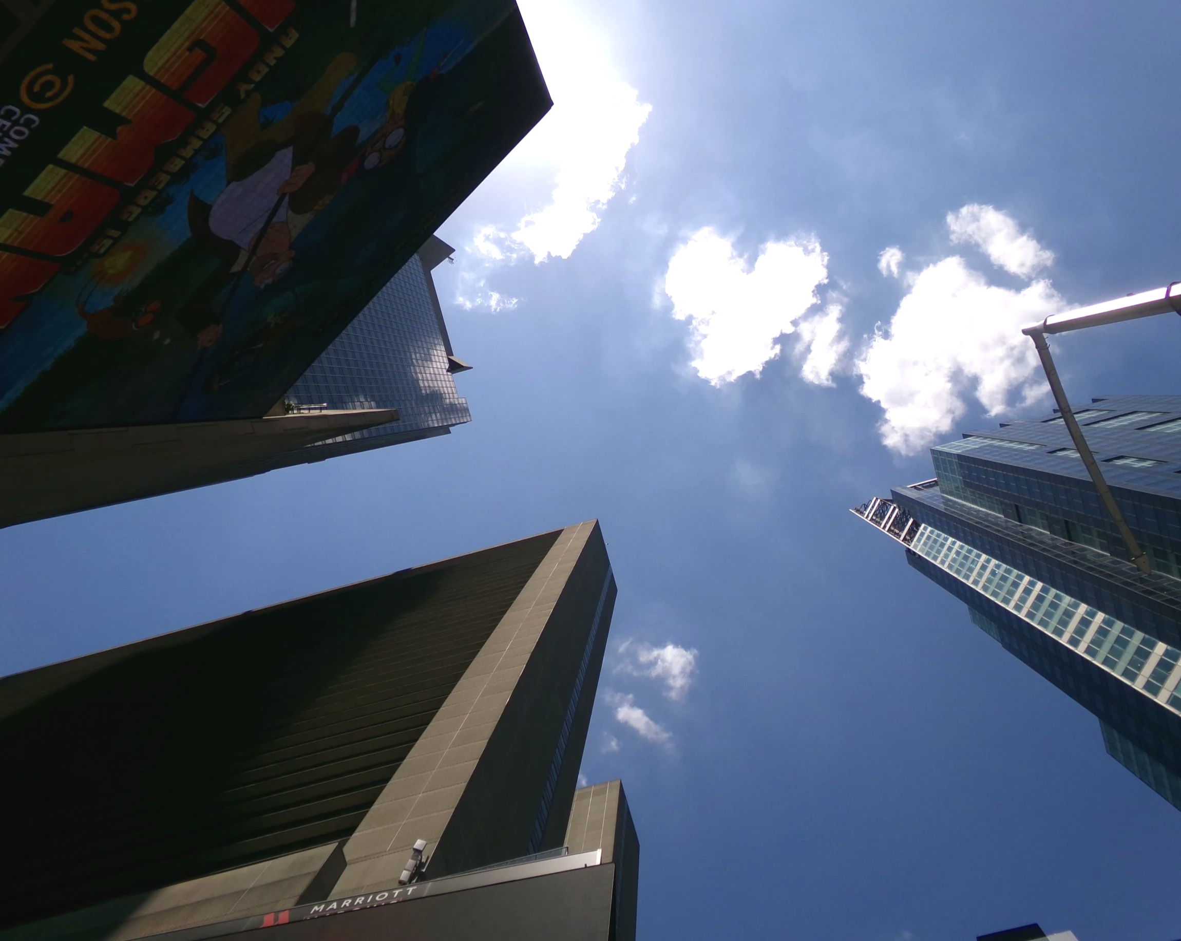 Looking up at tall modern buildings in a city, with a bright partly cloudy sky overhead.