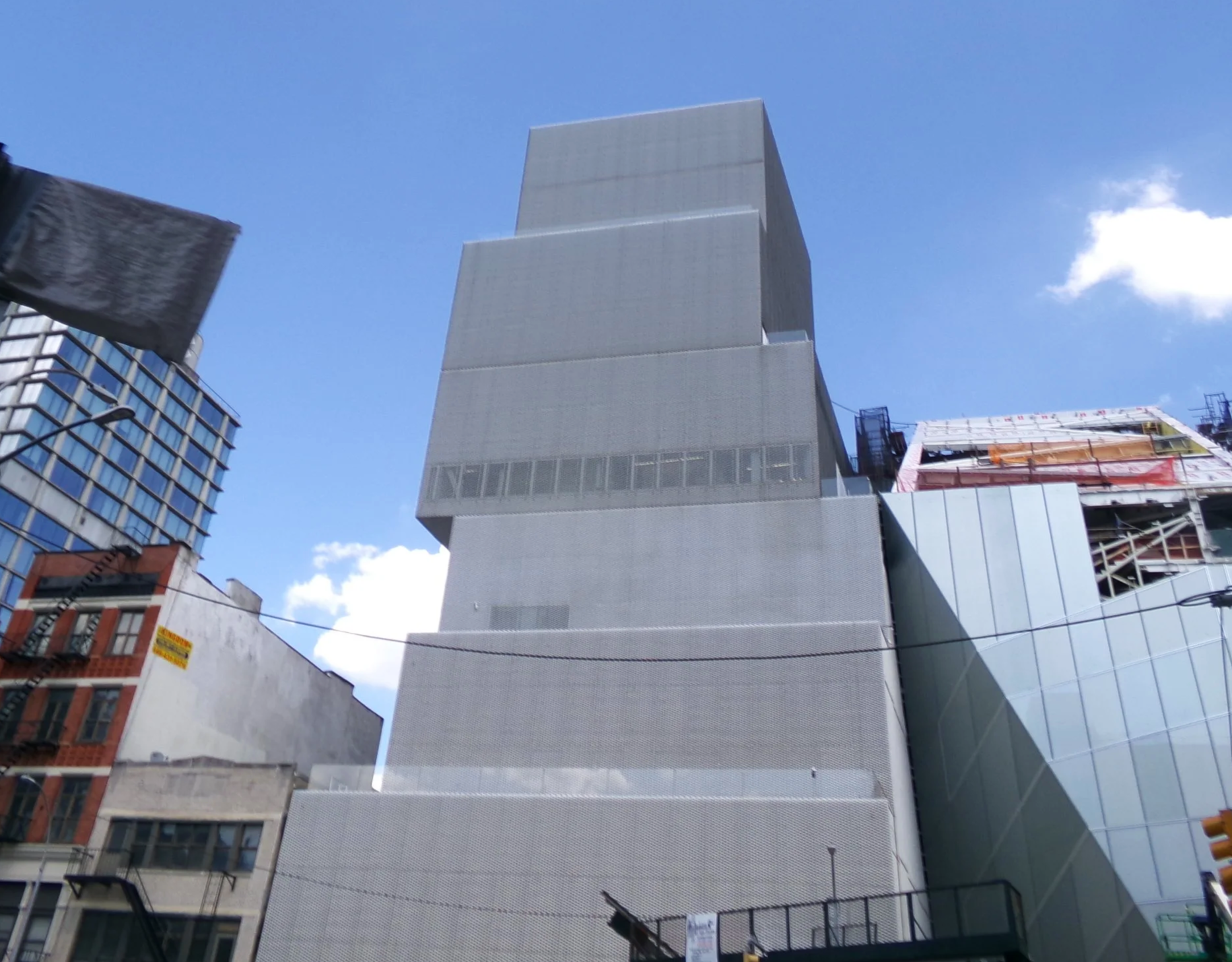A city street view looking up at tall modern buildings with a clear blue sky and some clouds, including a distinctive, stepped concrete building.