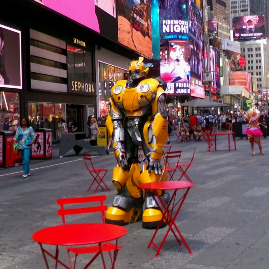 Times Square in New York City with digital billboards, red chairs and tables, a yellow robot statue, and a few pedestrians walking around on a sunny day.