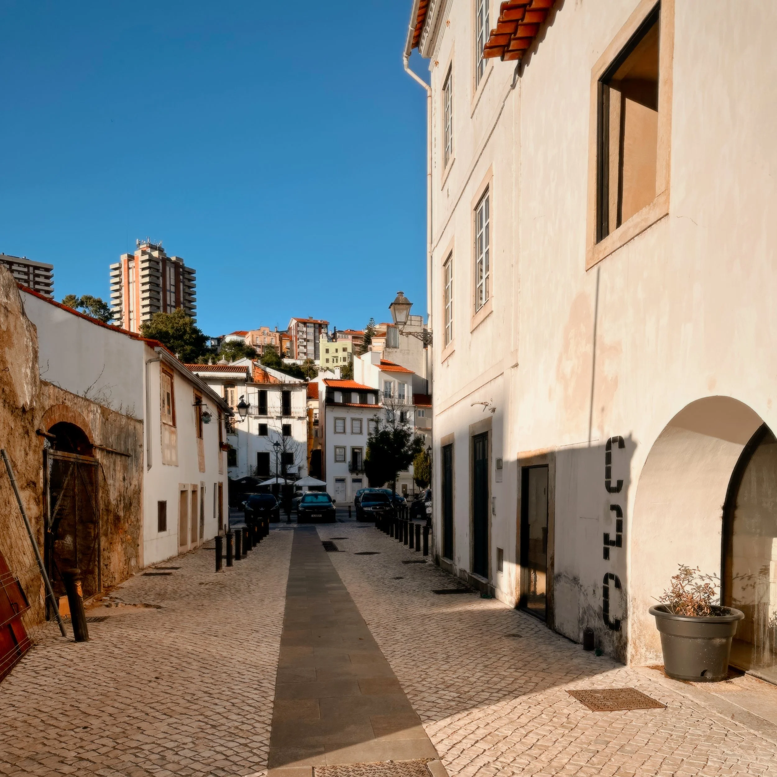 A narrow cobblestone street in a European city with white buildings on both sides. The street has a central paved section and black bollards on either side. In the background, modern high-rise buildings are visible under a clear blue sky.