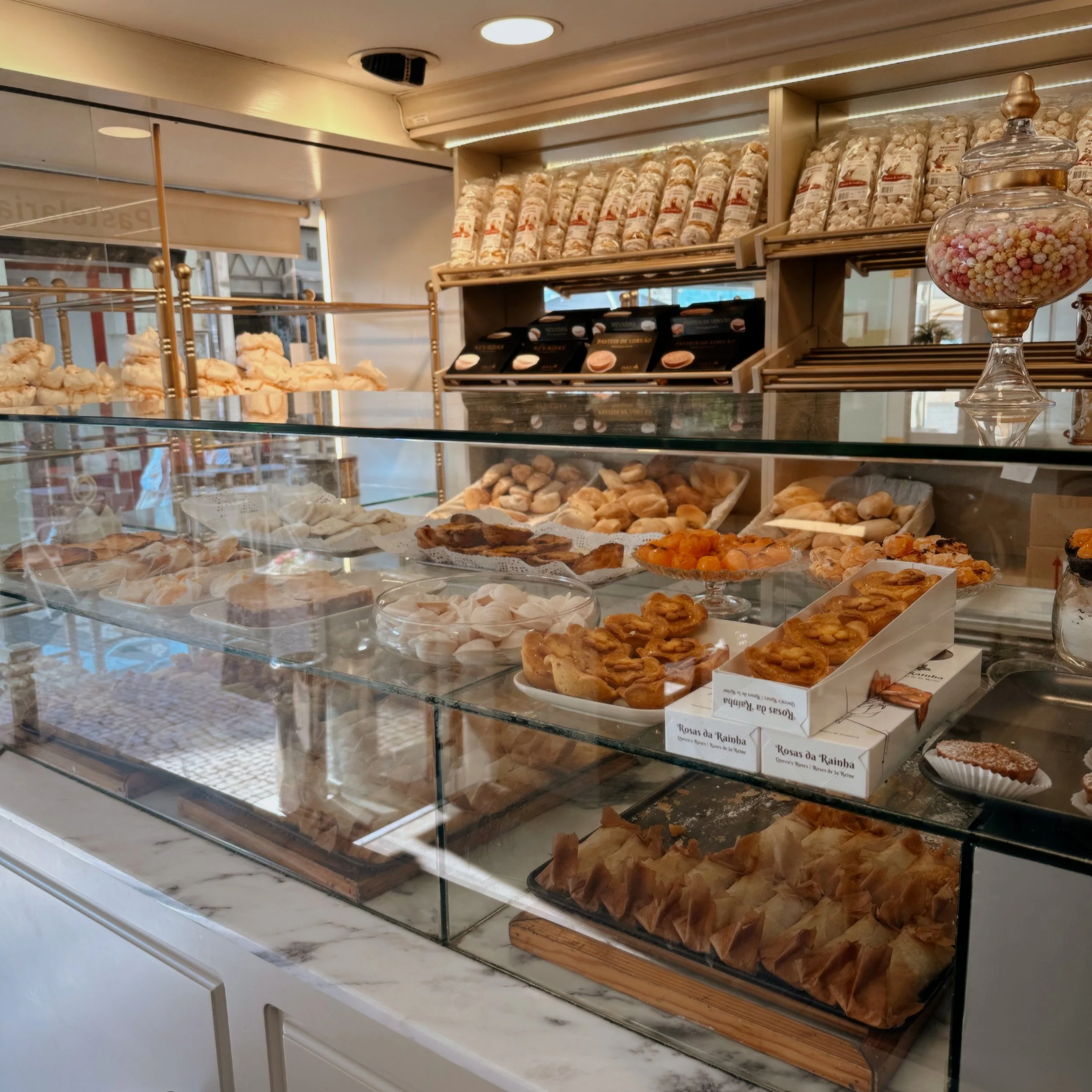 Display case with assorted pastries and baked goods, shelves with packaged sweets, and a jar filled with colorful round candies inside a bakery or café.