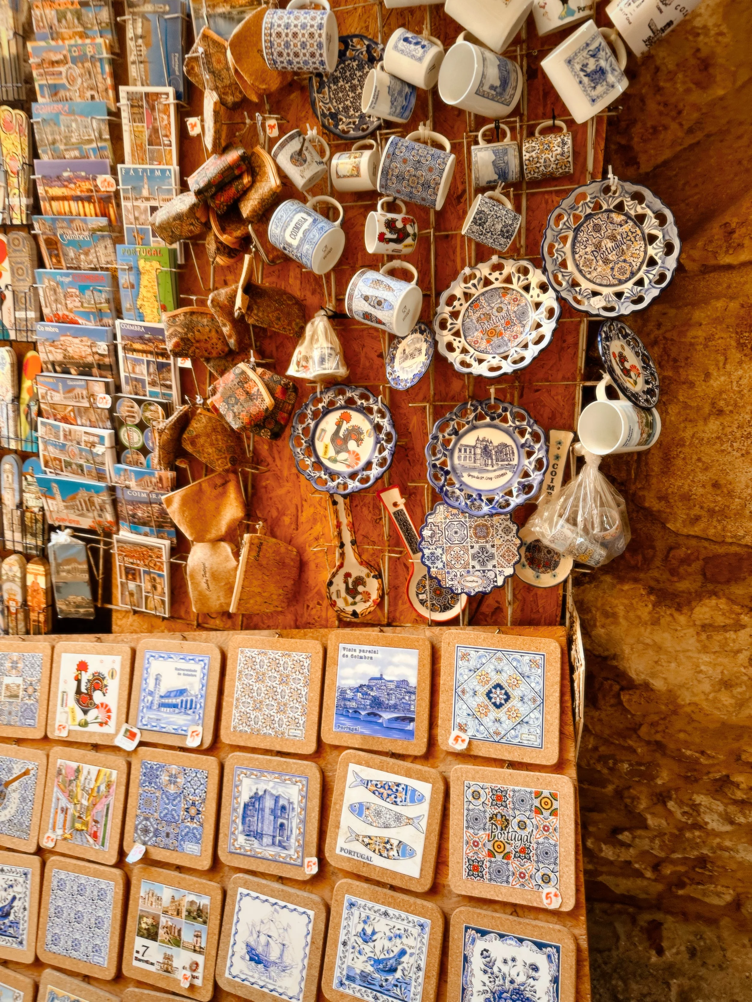 A display of souvenir ceramic mugs, plates, and tile coasters featuring Portuguese motifs and landmarks, with postcards of Portugal in the background.