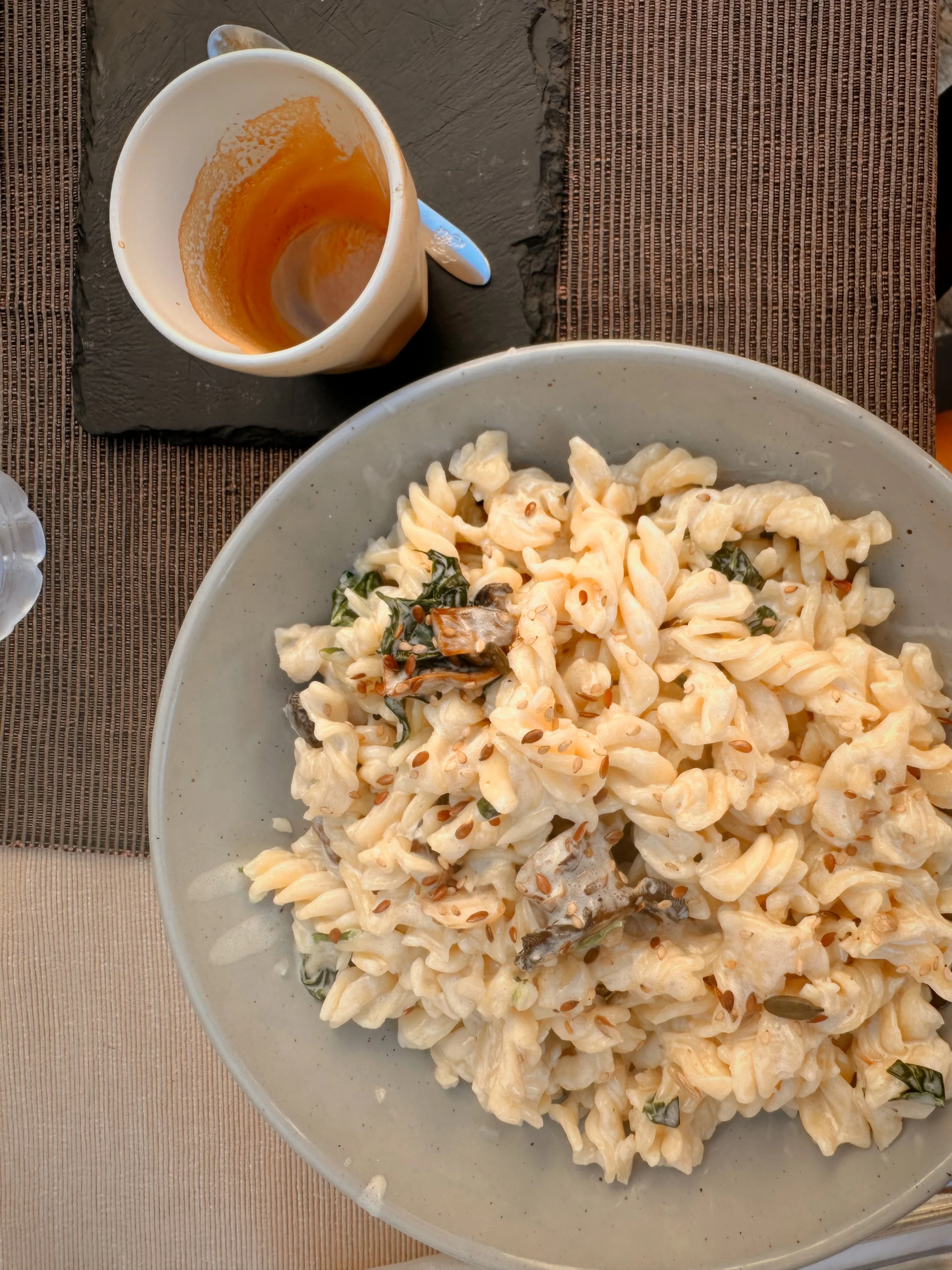 A plate of creamy pasta with green herbs and seeds, next to a cup with leftover sauce or coffee on a black slate coaster.