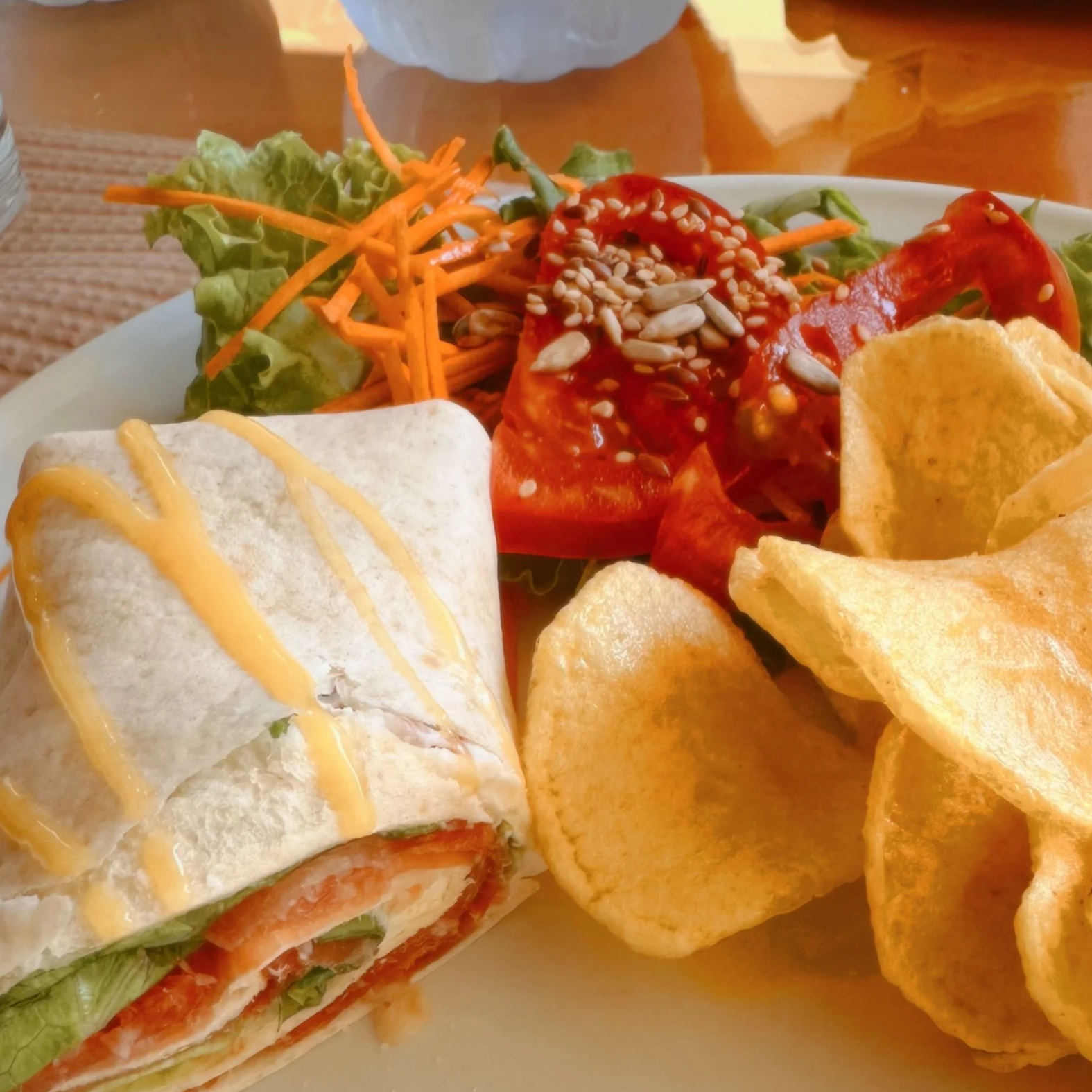A plate of food featuring a sandwich wrap with mustard, lettuce, tomatoes, and possibly other ingredients, accompanied by potato chips, on a dining table.