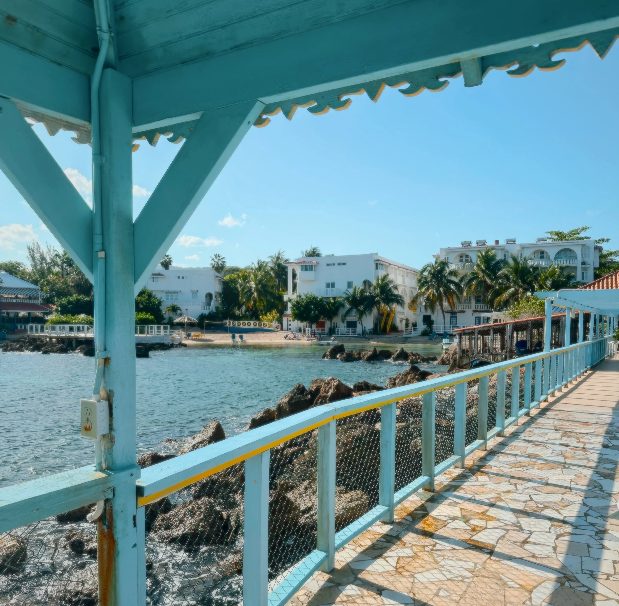 View from under a teal-colored pier over the water, showing rocks, a walkway, and residential buildings with palm trees and blue sky in the background.