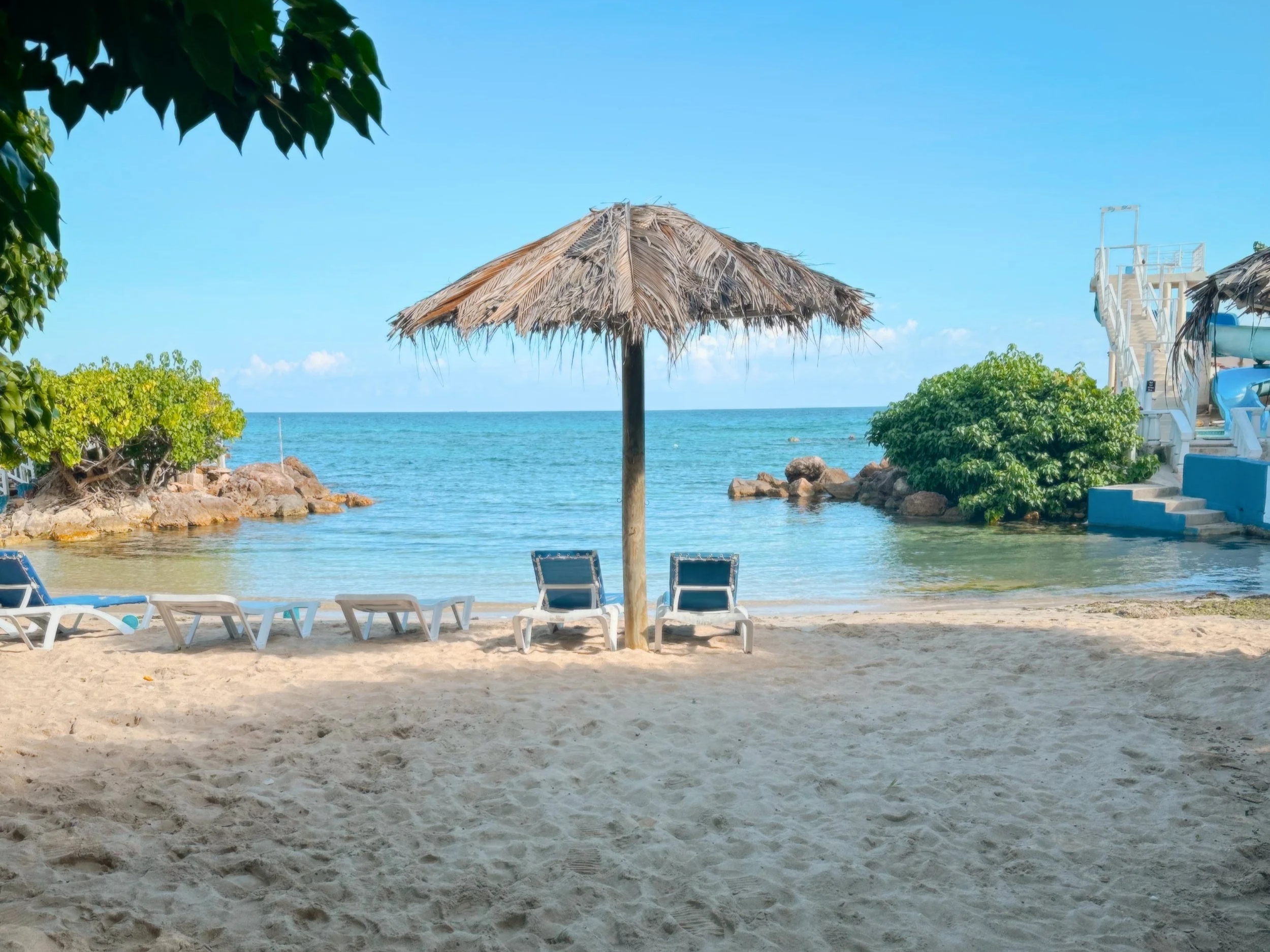 A beach scene with a straw umbrella, two chairs, and steps leading to an upper deck, with the ocean in the background.