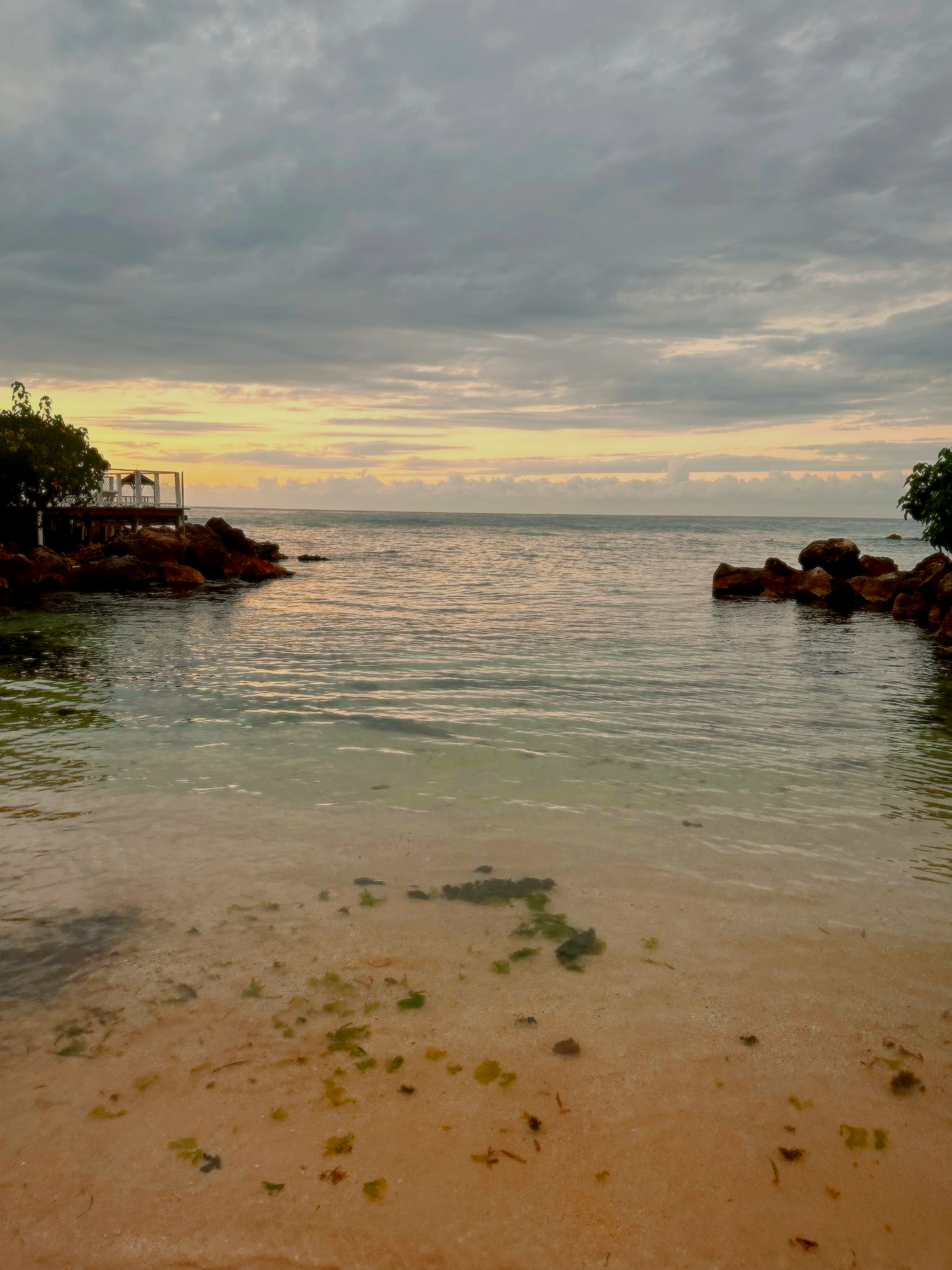 A beach scene with a sandy shore, rocks in the water, and a cloudy sky at sunset.