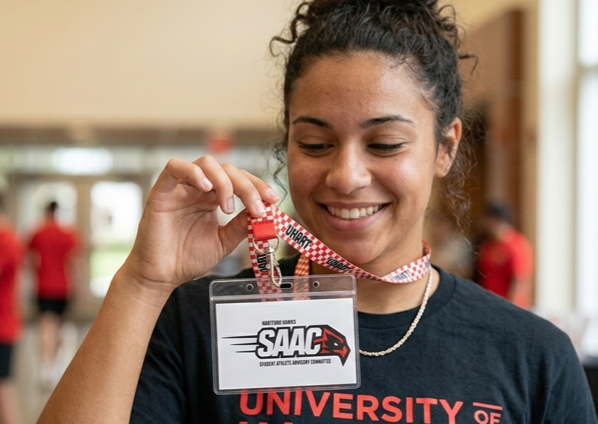 Young woman with curly hair smiling and holding a SAAC badge with a lanyard, in a communal indoor space with blurred people in the background.