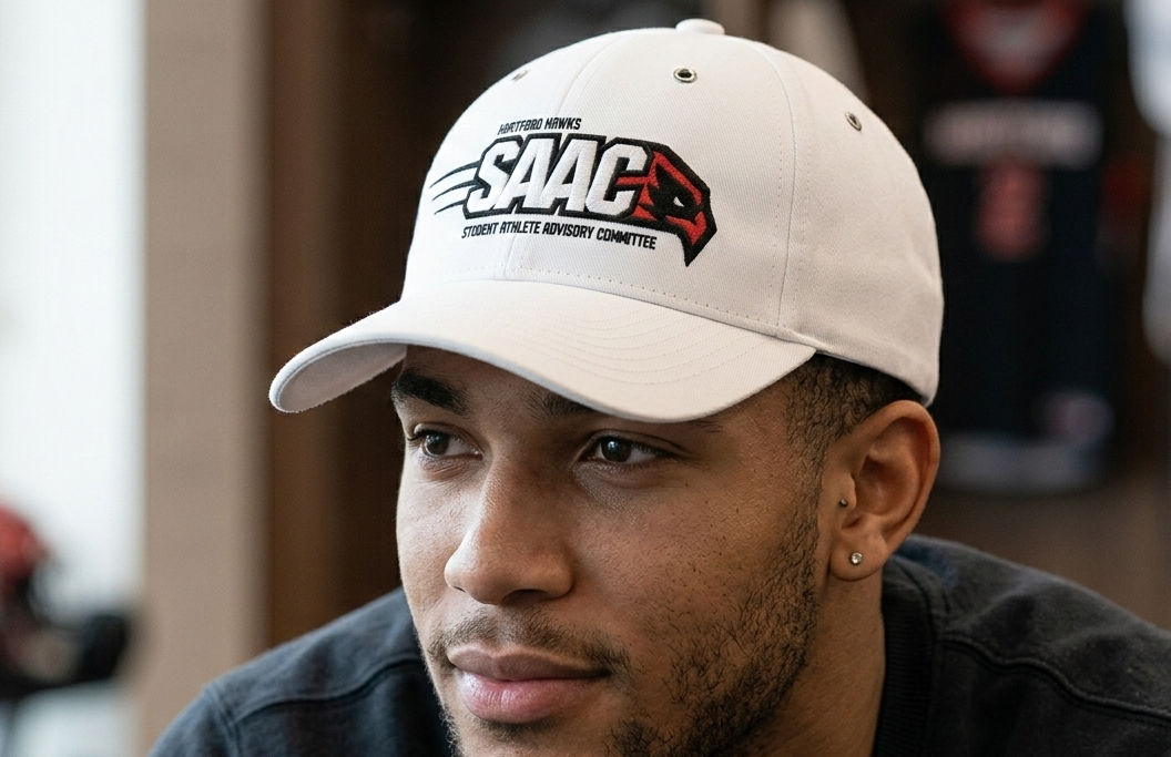 A young man wearing a white baseball cap with SAAC logo and text, and a black shirt, sitting indoors.