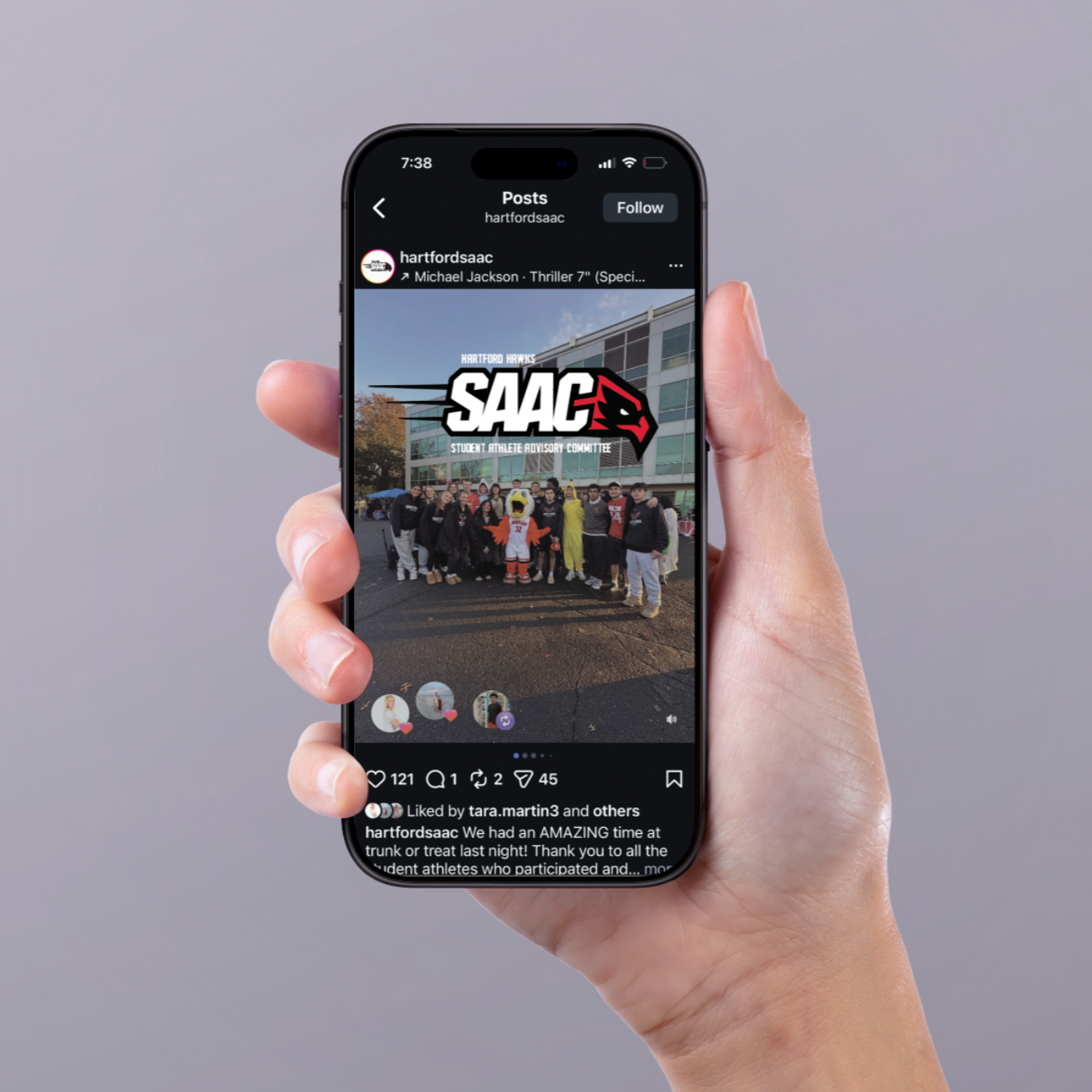 A hand holding a smartphone displaying an Instagram post with a group photo of people gathered outside a building, some in sports uniforms, with a mascot dressed as an eagle. The post features a large logo for "SAAC" and text indicating it is from th