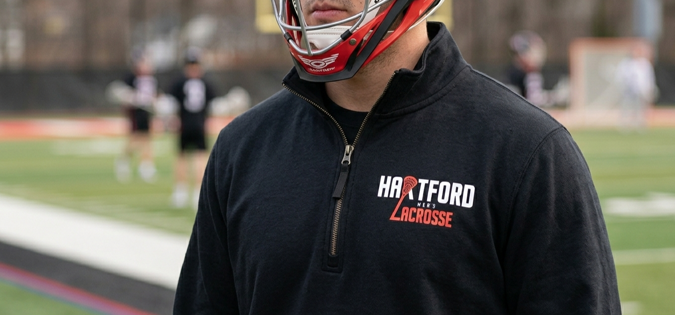 Close-up of a lacrosse coach or player wearing a black jacket with 'HARTFORD NER'S LACROSSE' logo, standing on a sports field with other players in the background.