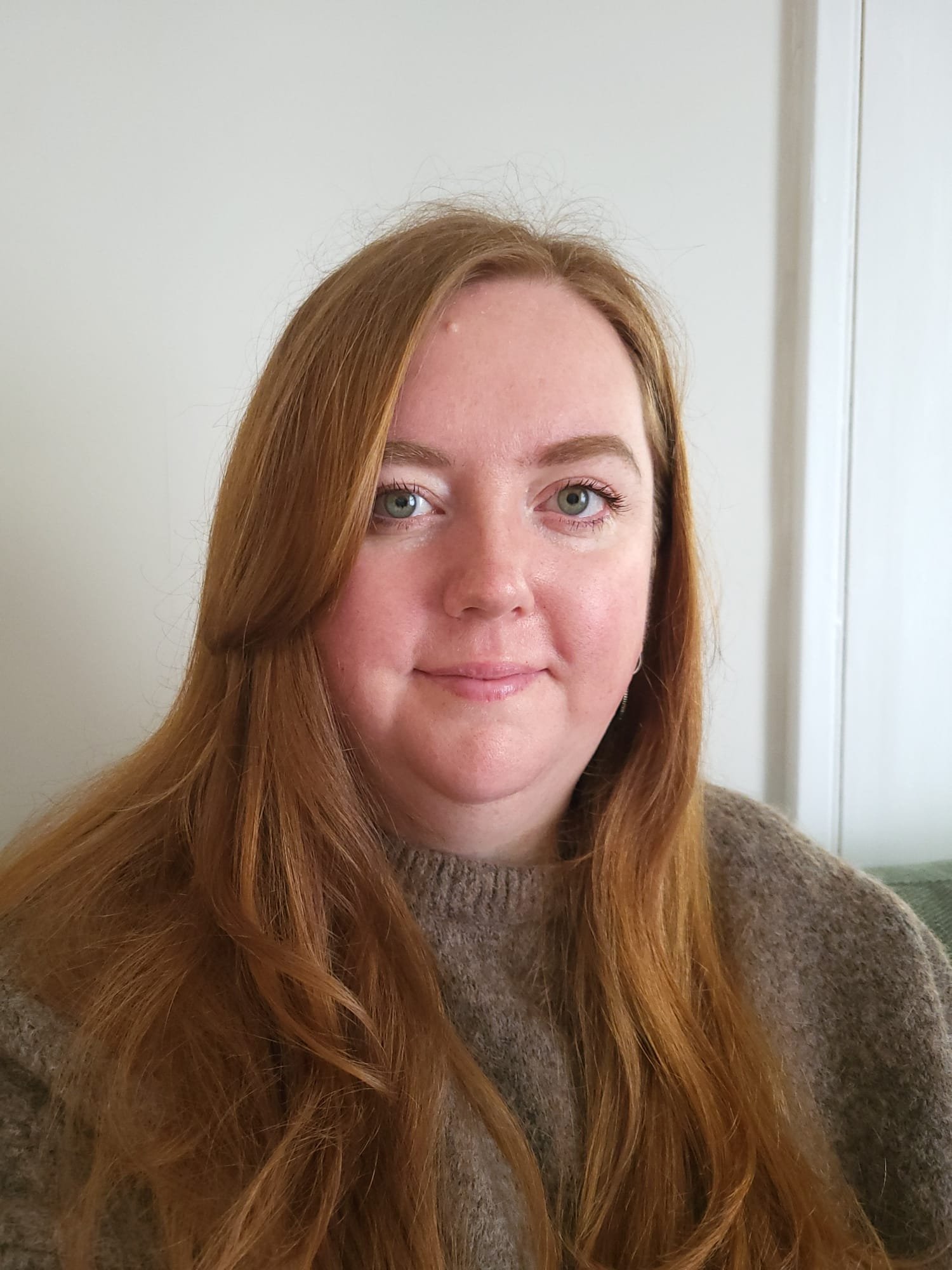A portrait of a young woman with long red hair and blue eyes, wearing a brown sweater, standing indoors with a white wall and a door in the background.