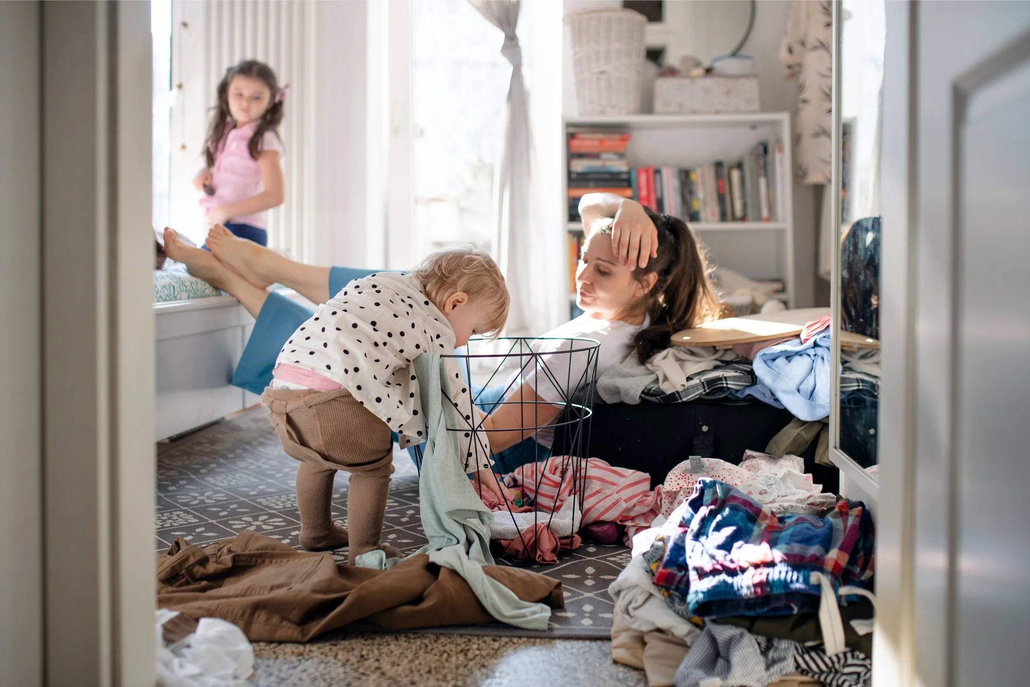 A woman lying on a bed, surrounded by piles of clothing, with two young girls in a messy bedroom, one standing and one leaning over a laundry basket.