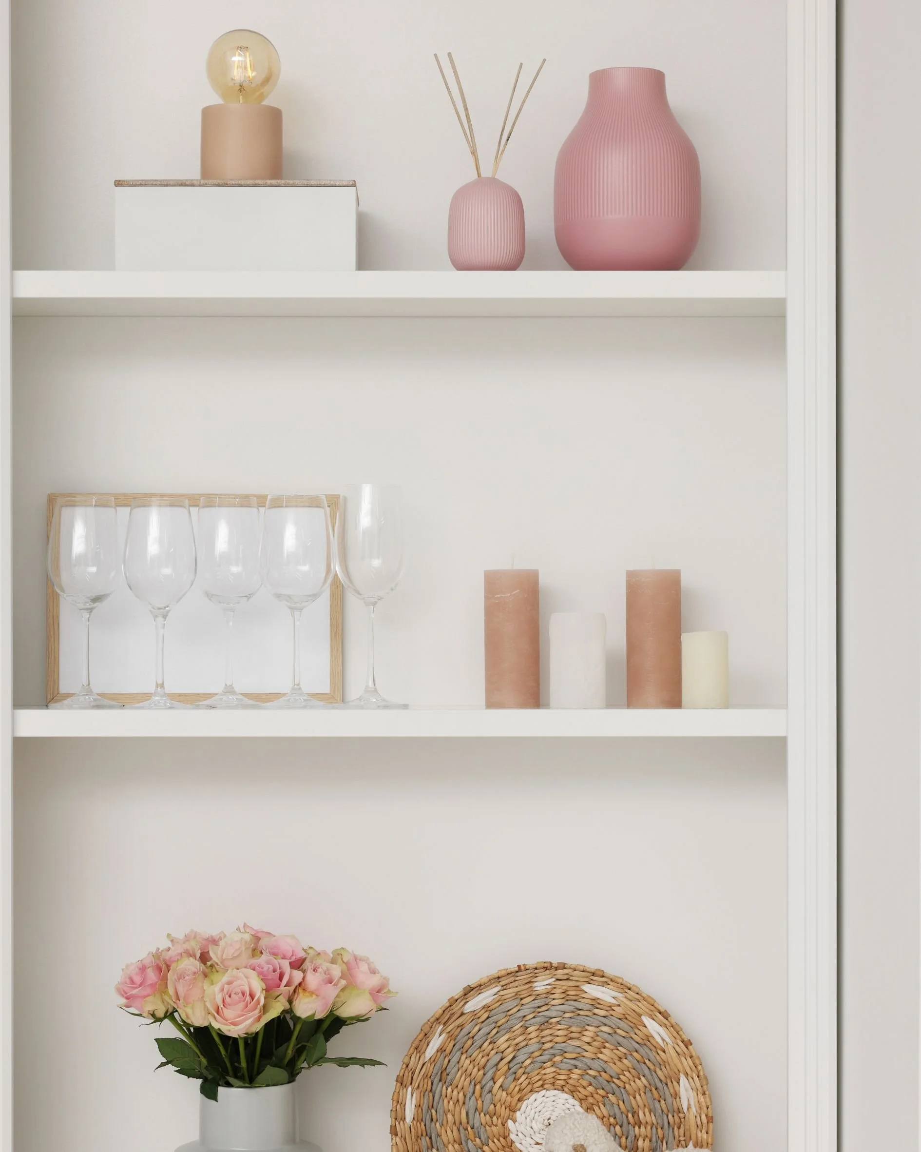 A white shelf displaying decorative items including pink vases, a lamp, glassware, candles, and a bouquet of pink flowers in a white vase.