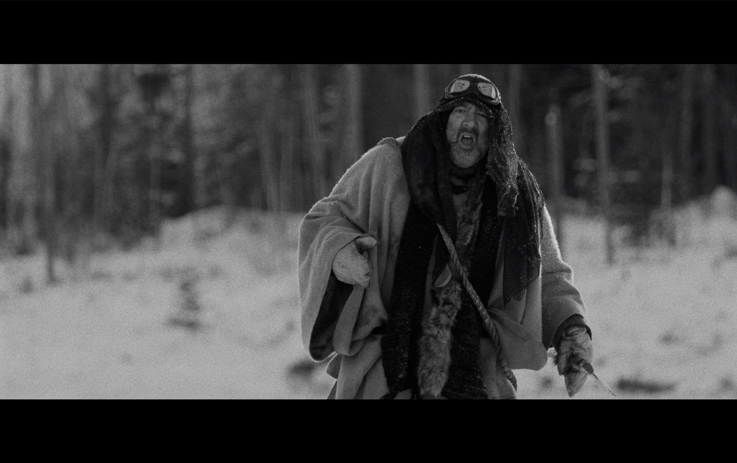 Homme avec un manteau lourd et des lunettes de ski, ayant une barbe grise, dans un paysage enneigé, l'air en colère ou surpris.