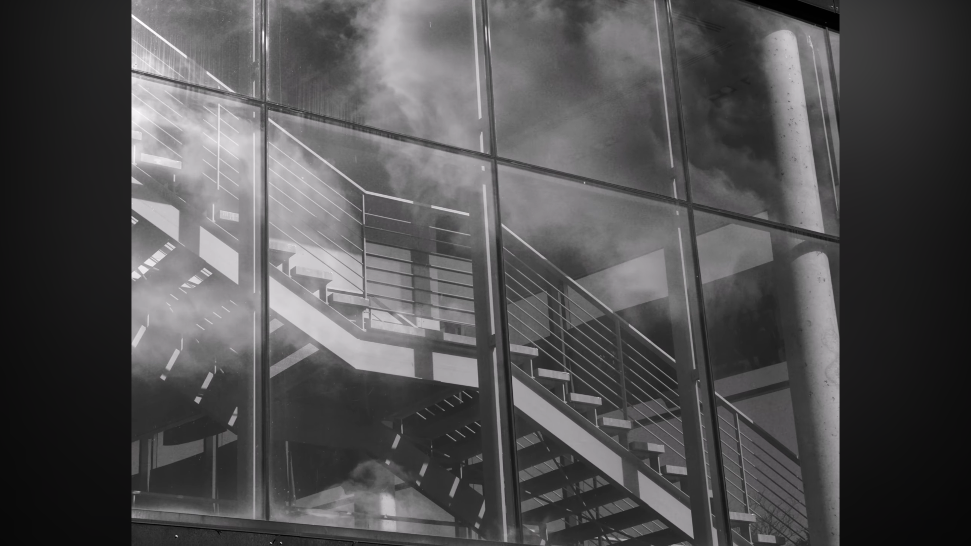 Escalier en métal visible à travers une façade en verre, avec des reflets de nuages dans le verre.