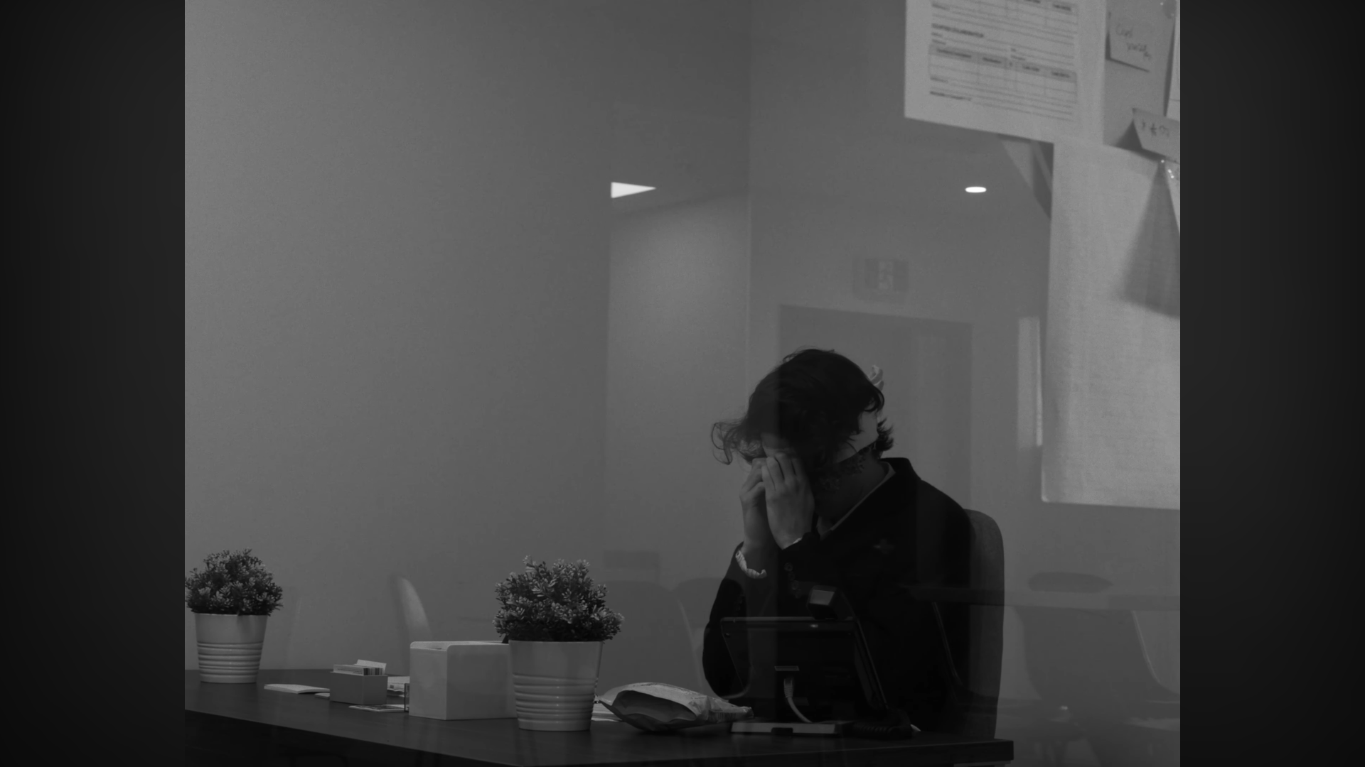 Une personne assise à une table vue à travers une vitre, avec des plantes en pot, des documents et un téléphone posé sur la table, en intérieur.