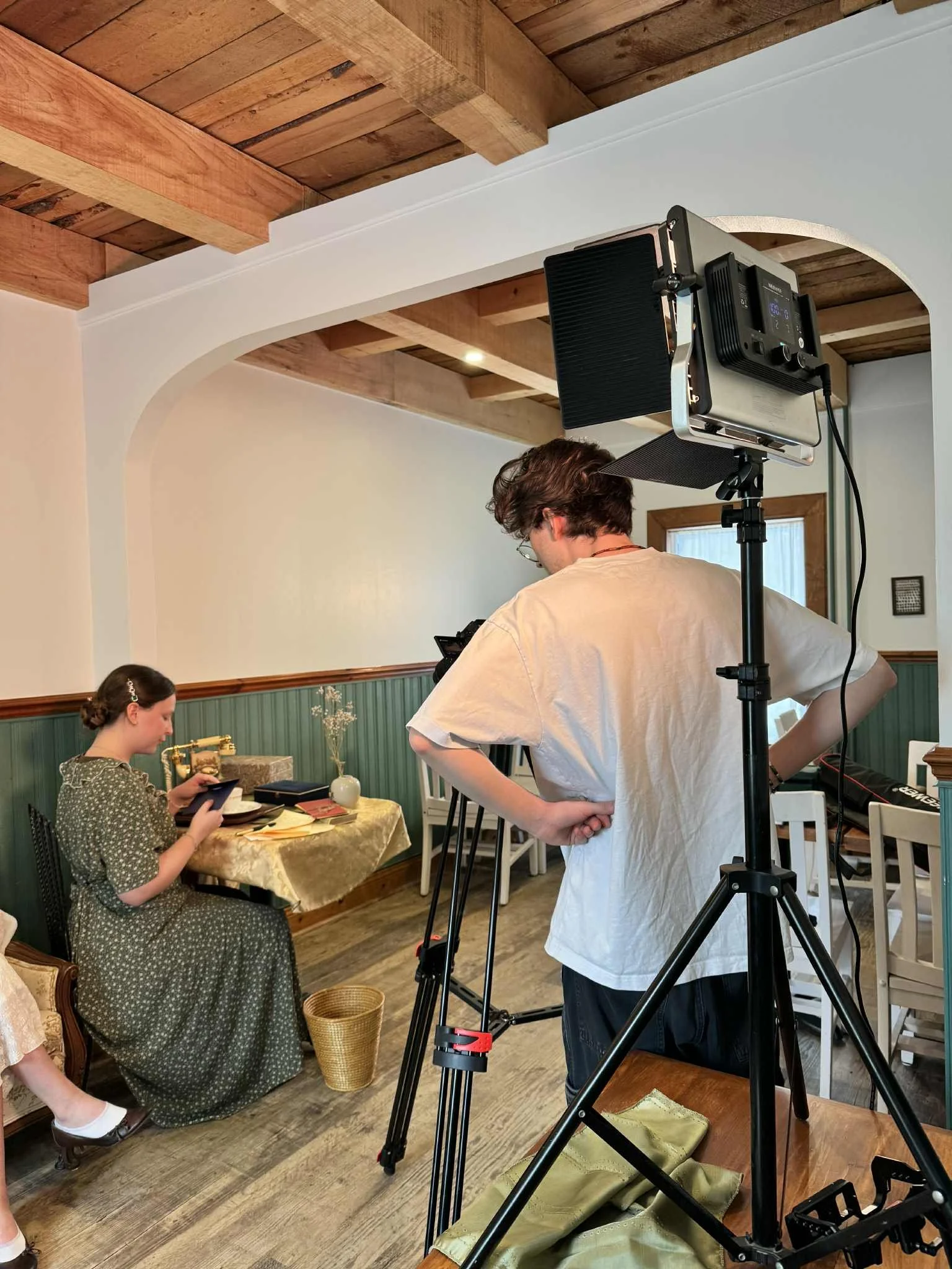 Une personne filme une scène dans une maison avec un éclairage professionnel, une jeune femme assise à une table de cuisine, lisant ou utilisant une tablette, décorée avec des vases et des objets anciens.