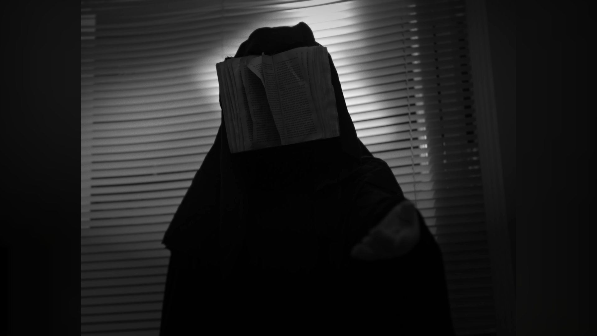 Une personne dont le visage est couvert, avec un journal sur la tête, dans une pièce sombre avec des stores à lamelles blanches en arrière-plan.