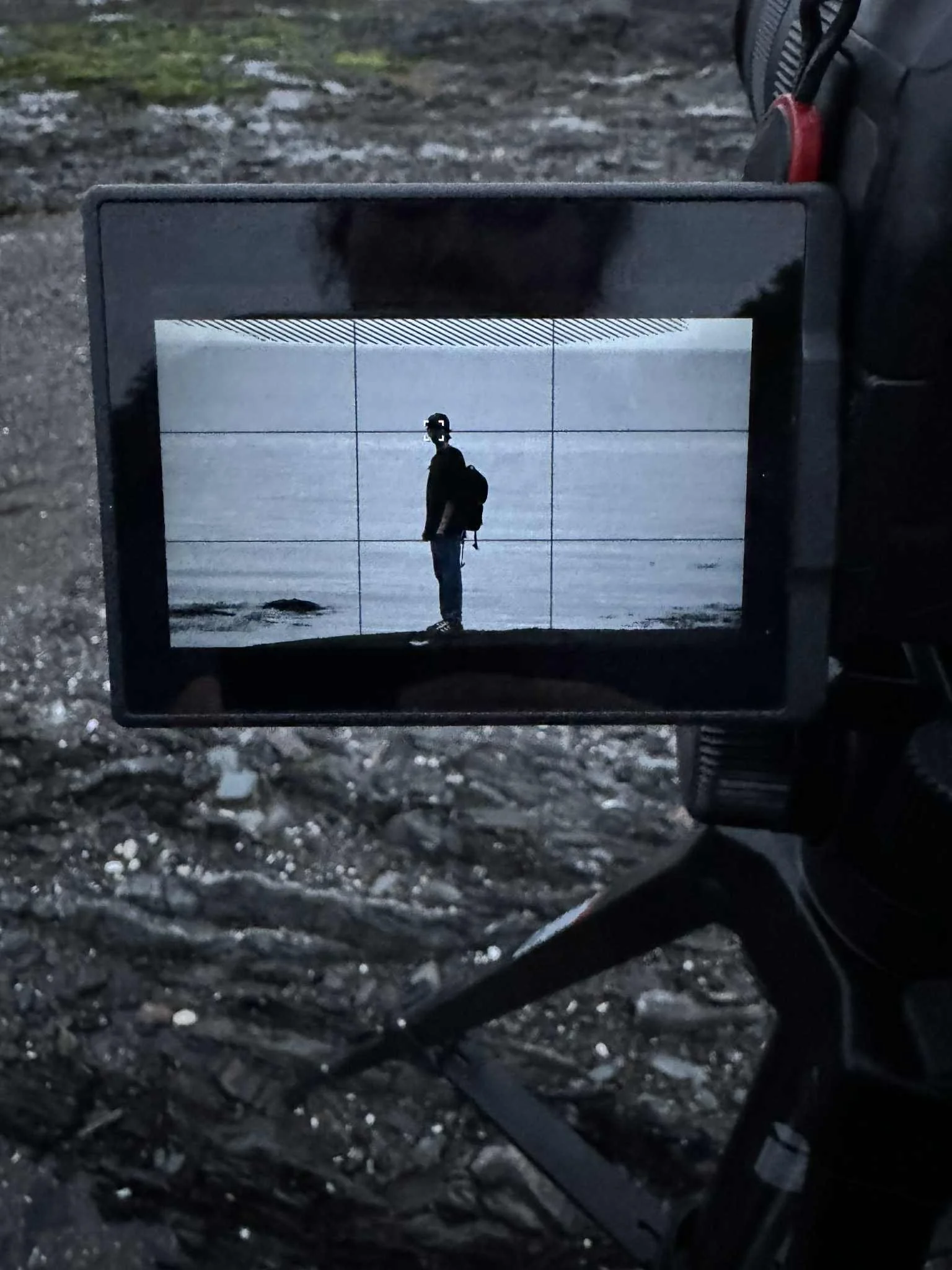 Un homme avec un sac à dos, vu à travers l'écran d'une caméra, se tient sur une plage ou une zone sablonneuse.
