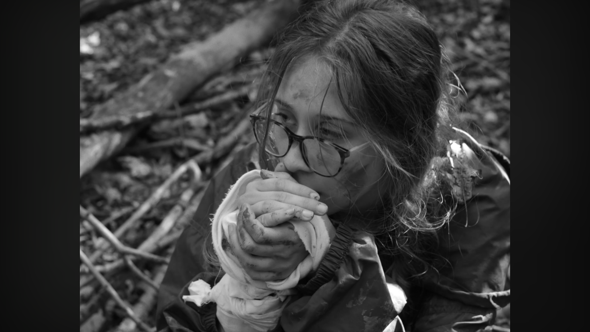 Une jeune femme portant des lunettes, dans un environnement forestier, tient sa main couverte de boue et de débris, semblant être dans une situation de sauvetage ou de survie.