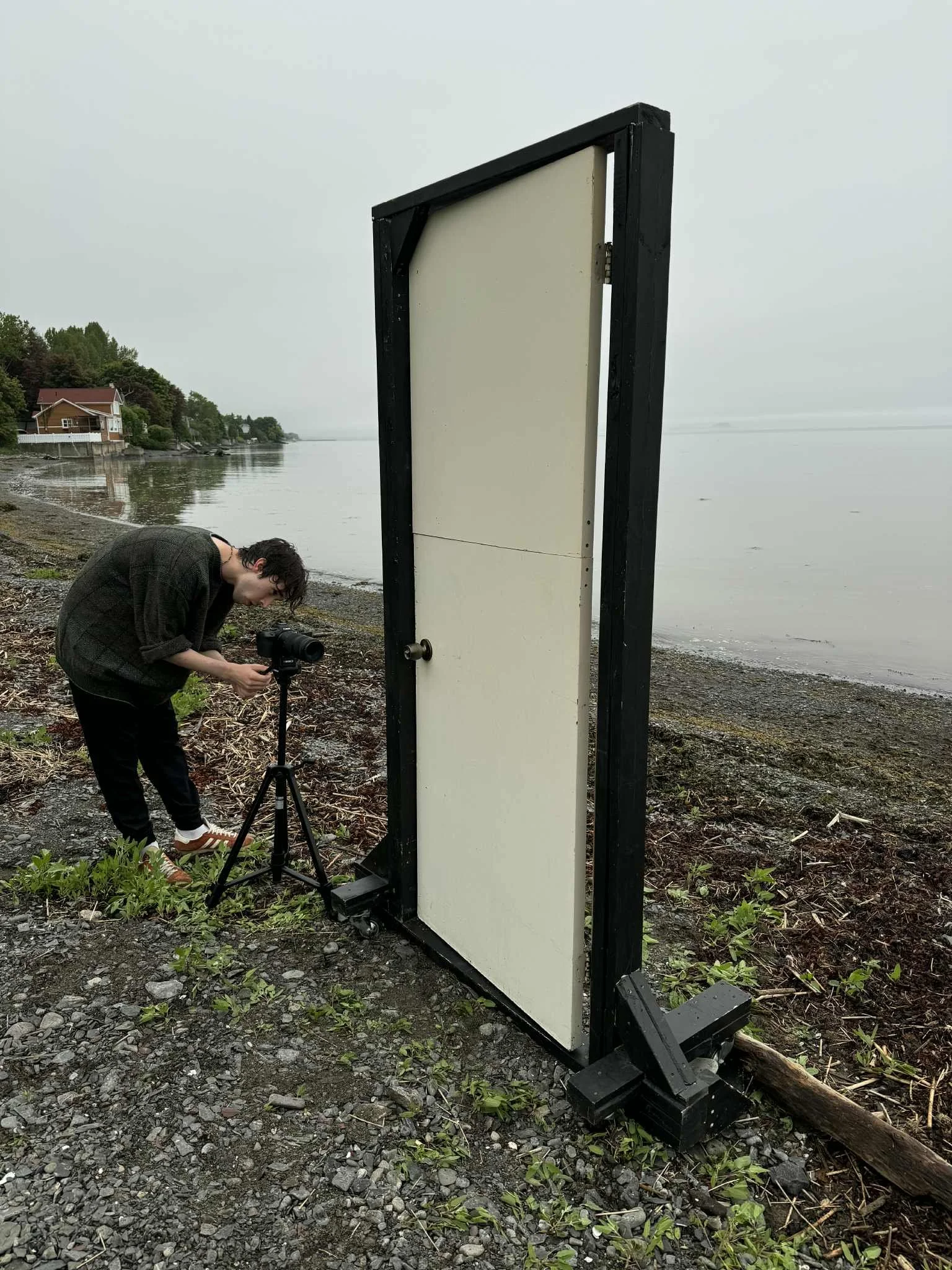 Une personne ajuste un appareil photo sur un trépied près de l'eau sur une plage rocheuse, à côté d'une structure noire et blanche sur la côte, par un temps nuageux.