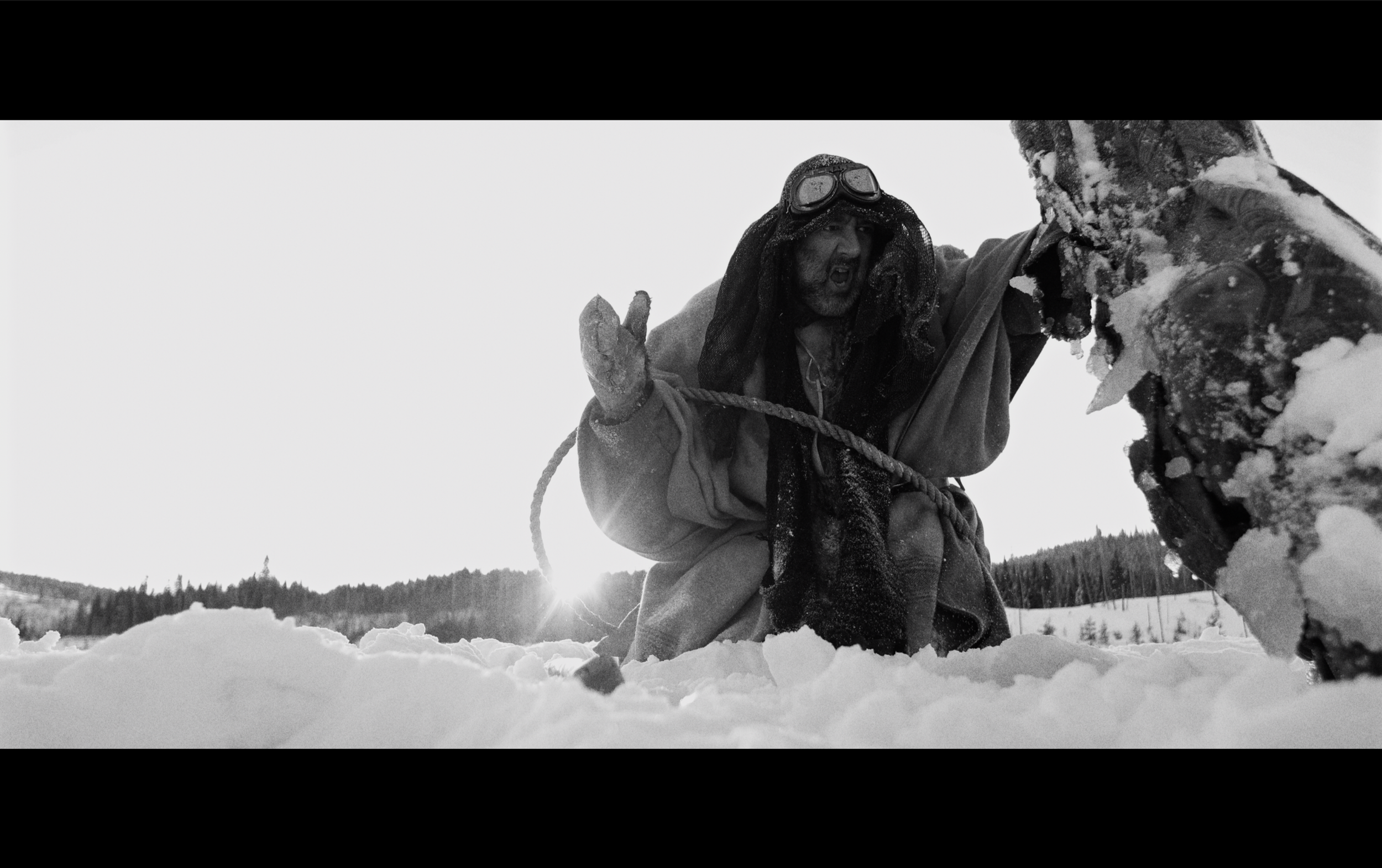 Un homme habillé en explorateur, avec des lunettes de protection, aide une autre personne dans la neige en utilisant une corde, dans un paysage enneigé.