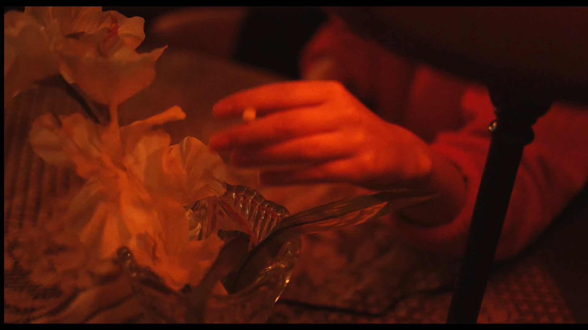 Une main tenant une cigarette dans un environnement sombre avec une lumière rouge/orange, des fleurs en arrière-plan et un objet en verre.