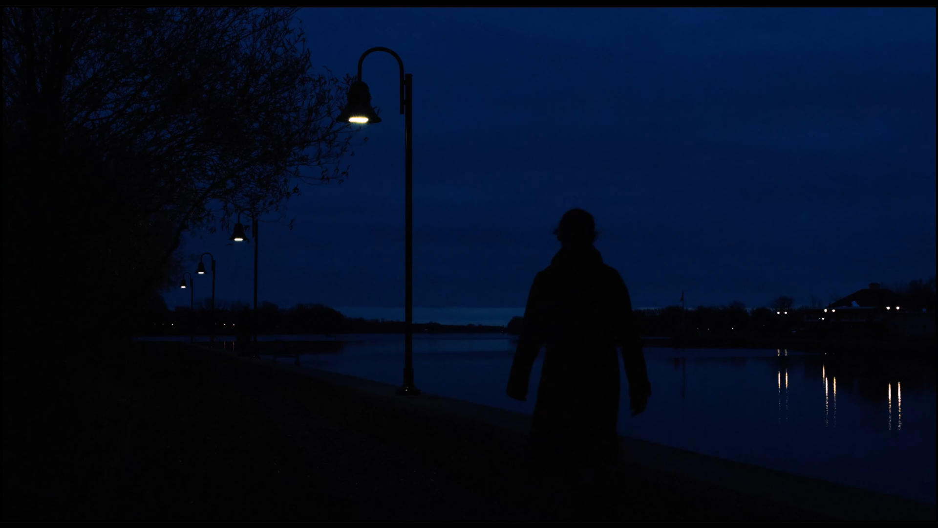 Silhouette d'une personne marchant le long d'une rivière de nuit, bordée de lampadaires allumés, avec des maisons éclairées en arrière-plan et un ciel sombre.