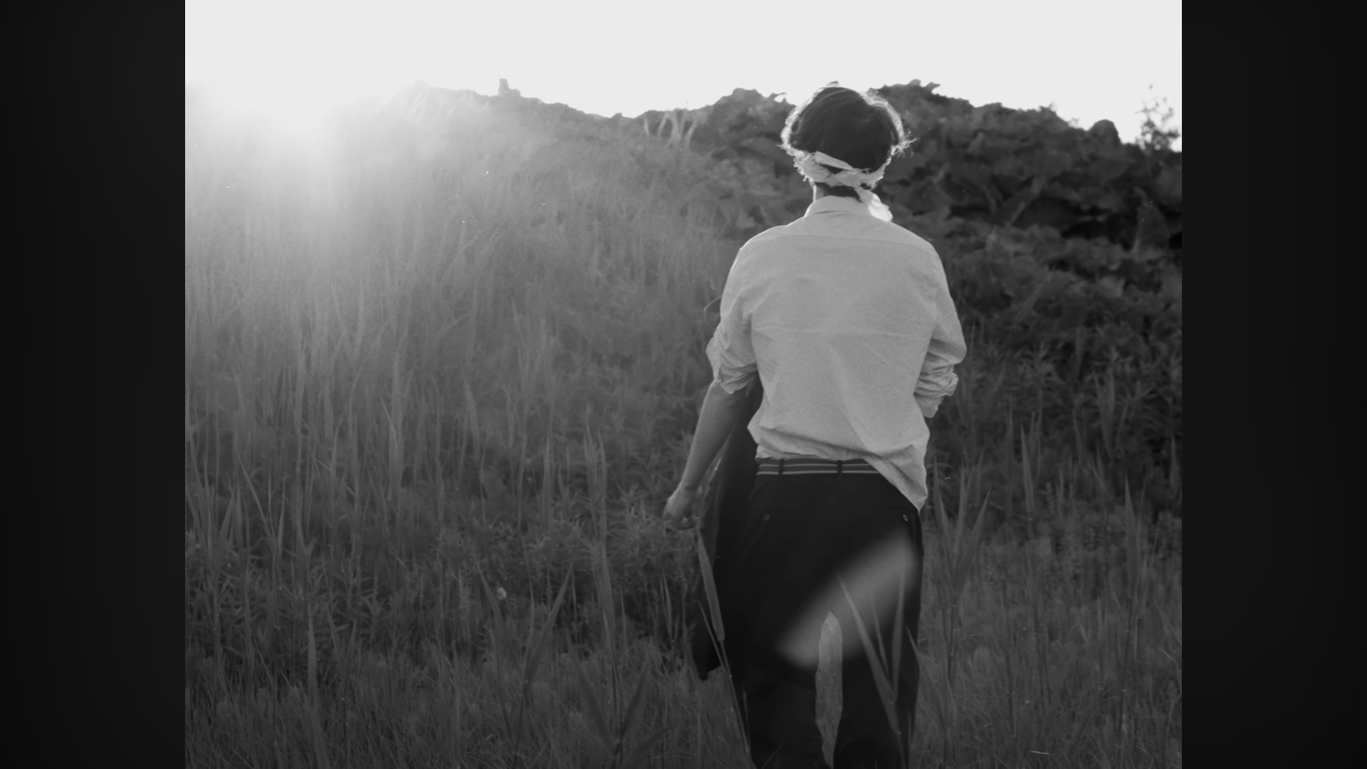 Une personne vue de derrière marchant dans un champ, avec le soleil couchant en arrière-plan, avec un style en noir et blanc.