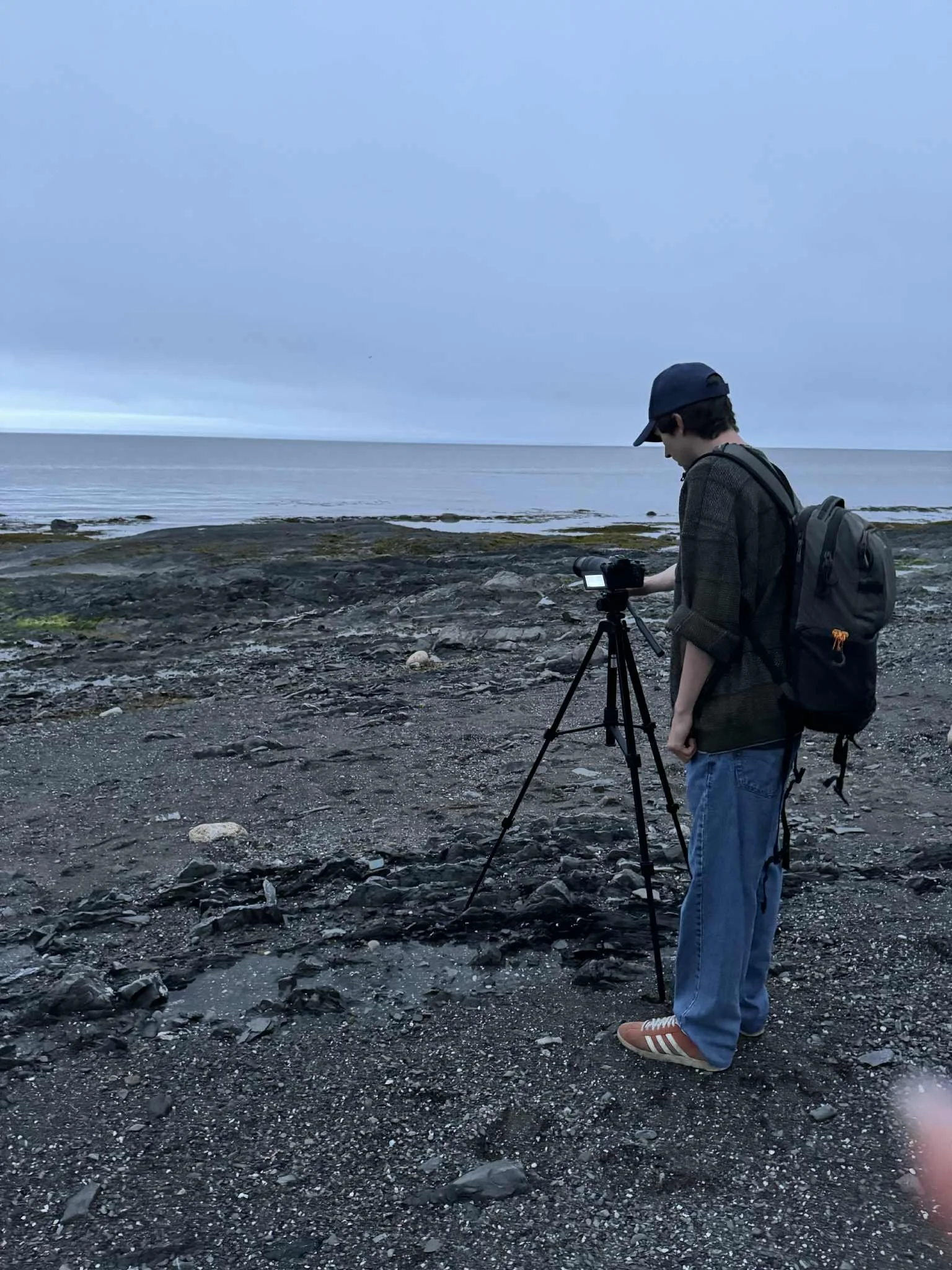 Jeune homme utilisant un appareil photo sur un trépied au bord de la mer, portant un sac à dos, une casquette et des vêtements décontractés.