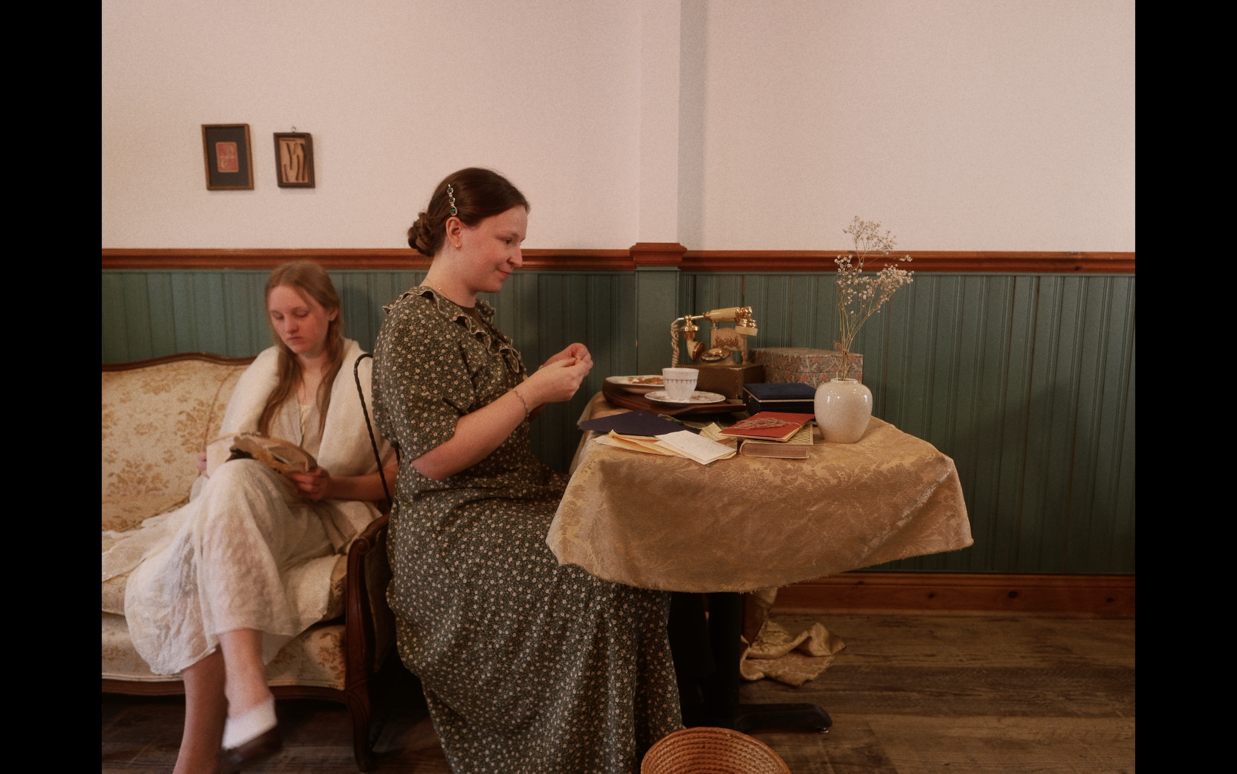 Deux femmes, l'une assise, l'autre debout, dans une pièce décorée de style vintage, avec un mobilier en bois, un vase avec des fleurs séchées, un téléphone ancien et des livres sur une table recouverte d'une nappe dorée