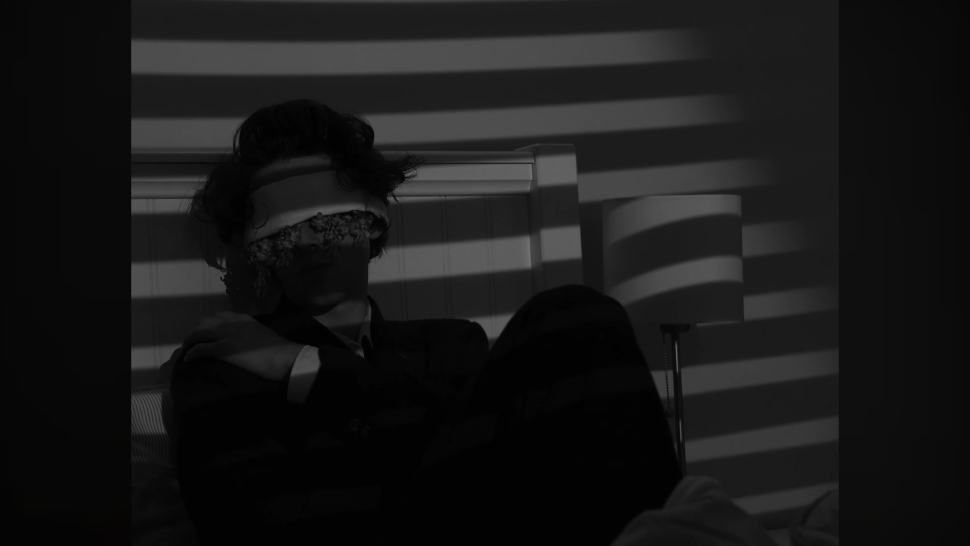 Une personne assise sur un lit, portant un bandeau sur les yeux, avec des rayures de lumière projetées sur le mur derrière elle.