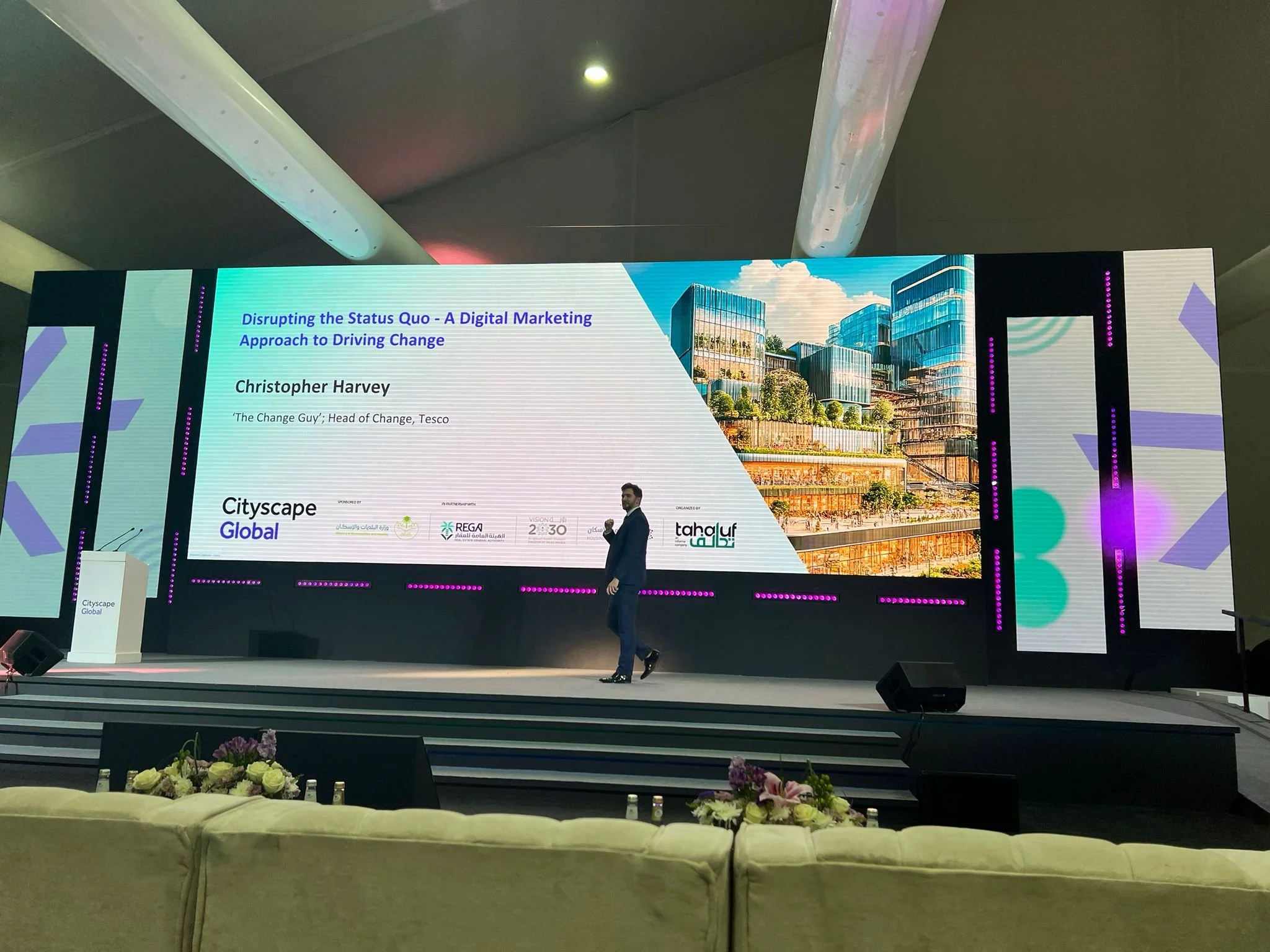 A presentation stage with a large screen displaying a slide titled "Disrupting the Status Quo - A Digital Marketing Approach to Driving Change" by Christopher Harvey, Head of Change at Tesco. The slide features an image of modern office buildings and logos of sponsors and partners. There is a man standing on the stage, and the foreground shows a row of flowers and a sofa.