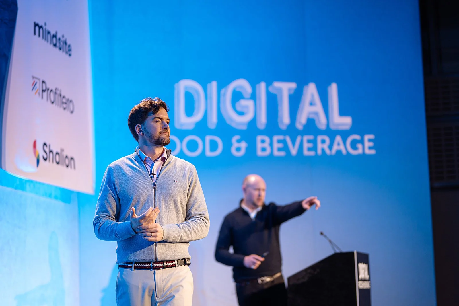 A man in a beige zip-up sweater giving a presentation at a conference, with another person in the background pointing at a large screen displaying the words 'Digital Food & Beverage' and logos of sponsors.