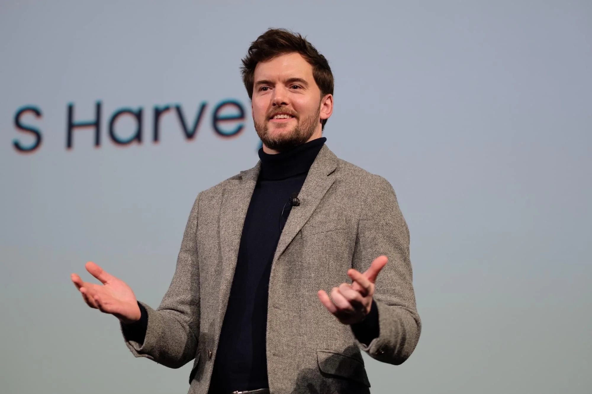 A man with brown hair and a beard wearing a grey blazer and black turtleneck speaking in front of a plain background.
