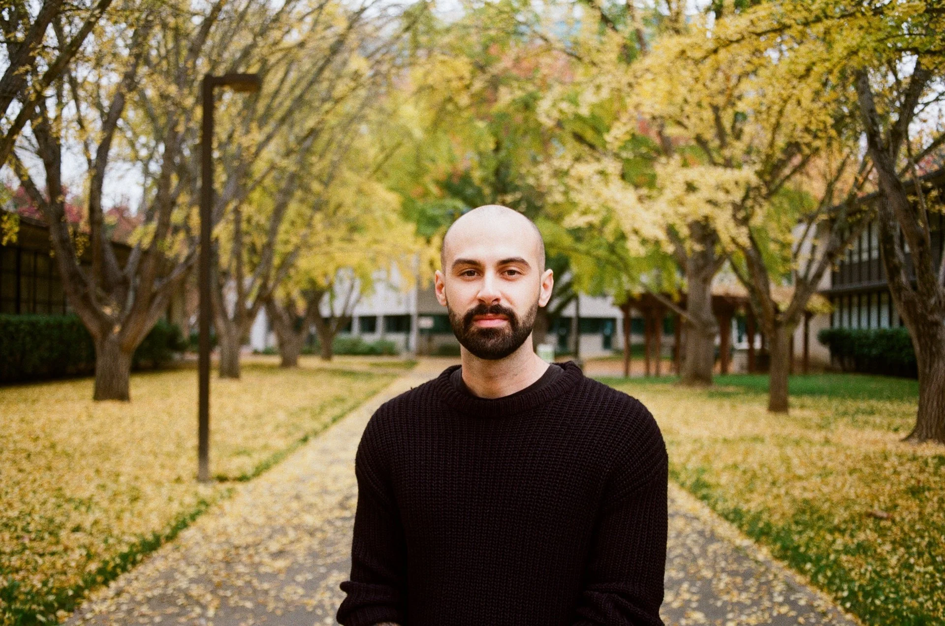 A man with a beard and bald head wearing a black sweater standing on a pathway in a park with fall foliage, yellow leaves on trees and on the ground