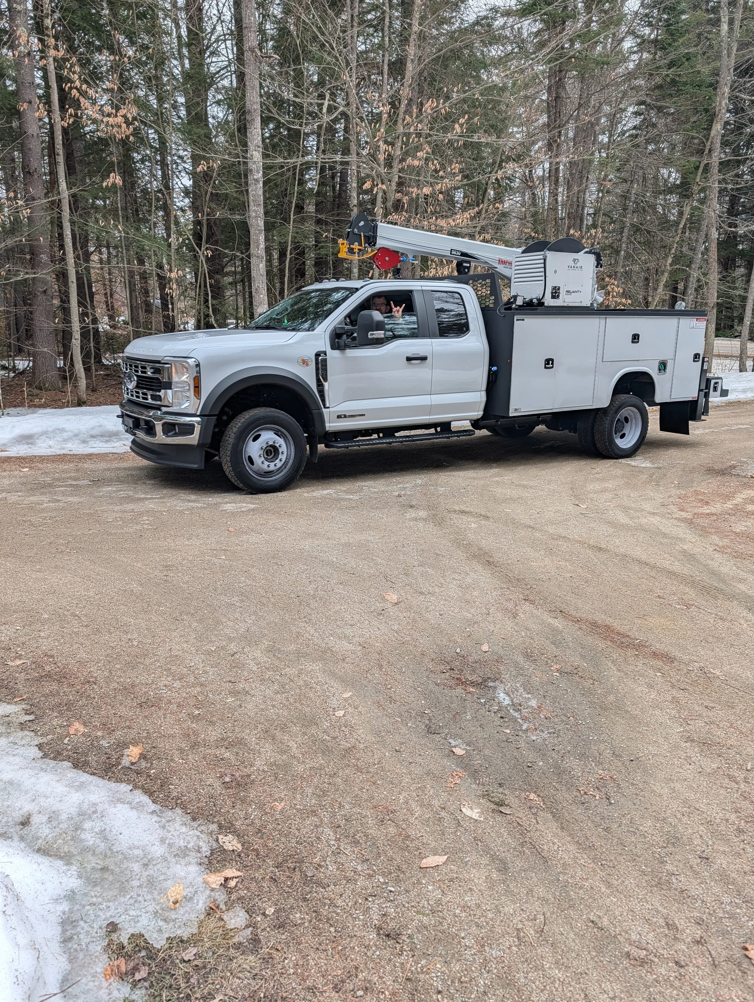 Silver utility truck with a mounted crane or lift, parked on a gravel road near a wooded area with some snow on the ground.  This truck is used for well maintenance.