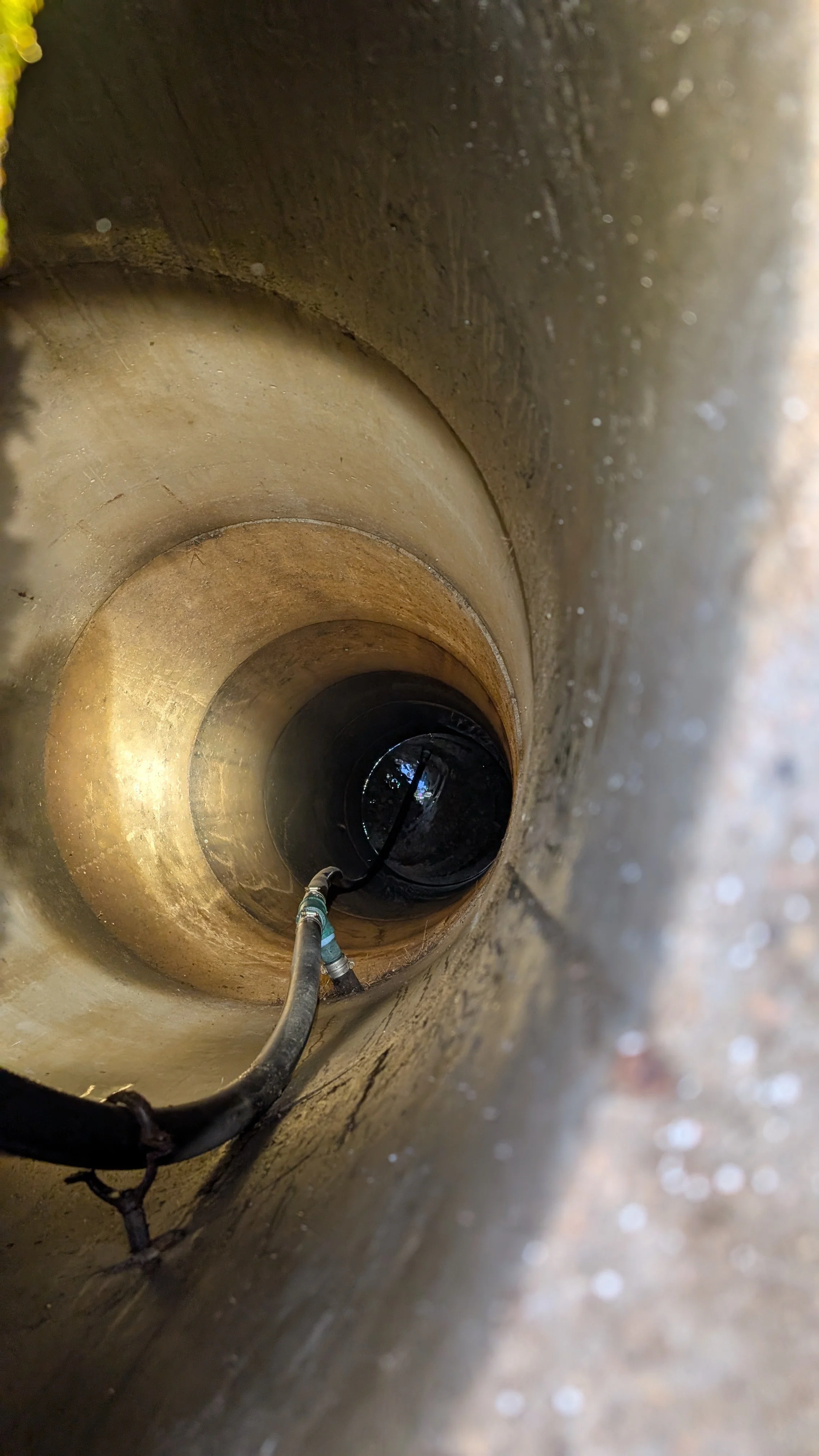 Inside a water well, looking down to the bottom of the wellwith a hose or cable extending towards the bottom of the pipe.  This shows the well pump inside of the well.