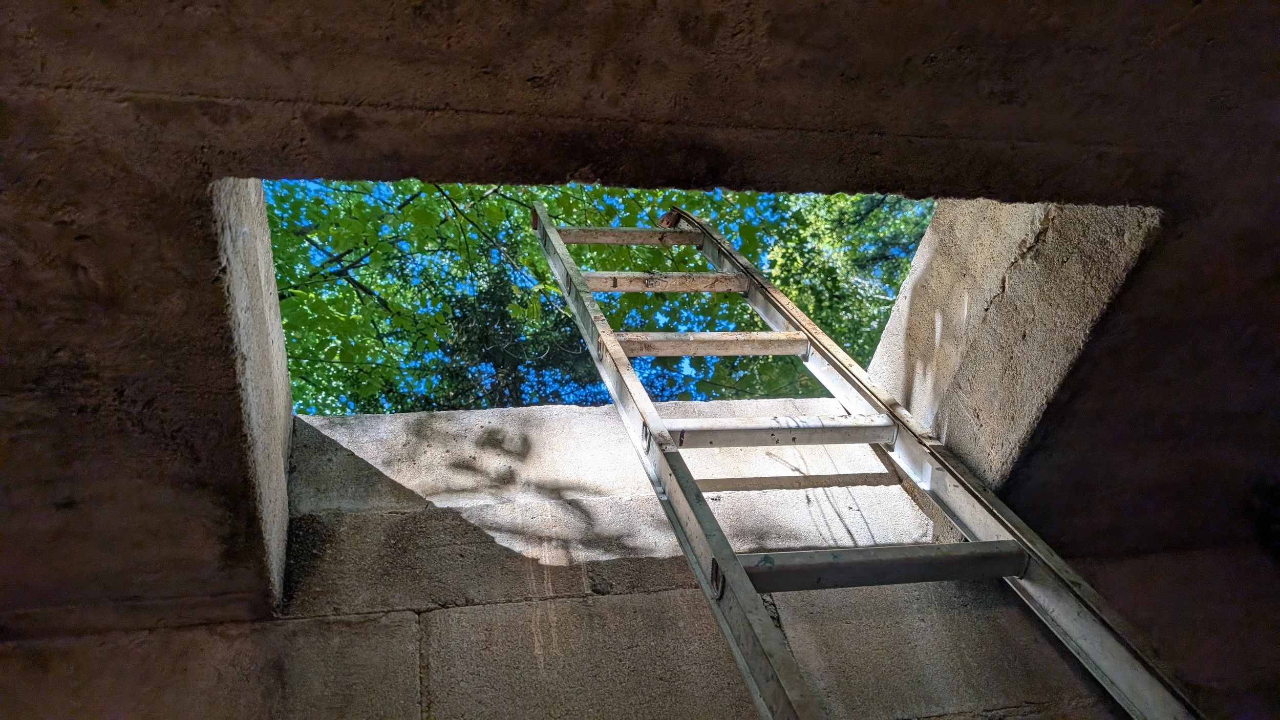 View looking up through an opening from a well indicating that a well is being repaired with green trees and blue sky visible through the opening.